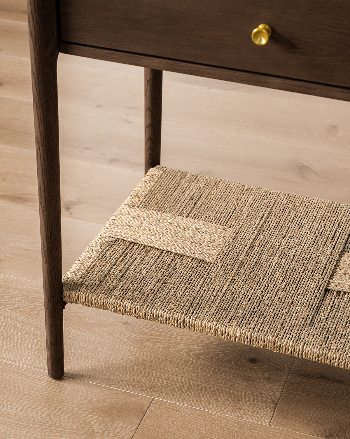 Close-up of the Dana Walnut Oak Console leg and woven shelf on a light wood floor. The console features a dark walnut oak finish and a drawer with elegant brass hardware, by Vendixs