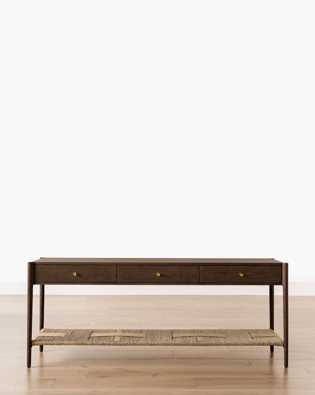 The Dana Walnut Oak Console, featuring three drawers, brass hardware, and a woven shelf, stands on a light wood floor against a plain white background, by Vendixs