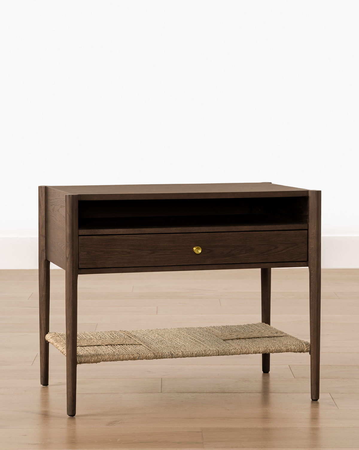 The Dana Walnut Oak Nightstand features slender legs, a wide drawer with a brass knob, an open shelf above, and a woven rattan bottom shelf. It is shown on a light wood floor against a white background, by Vendixs