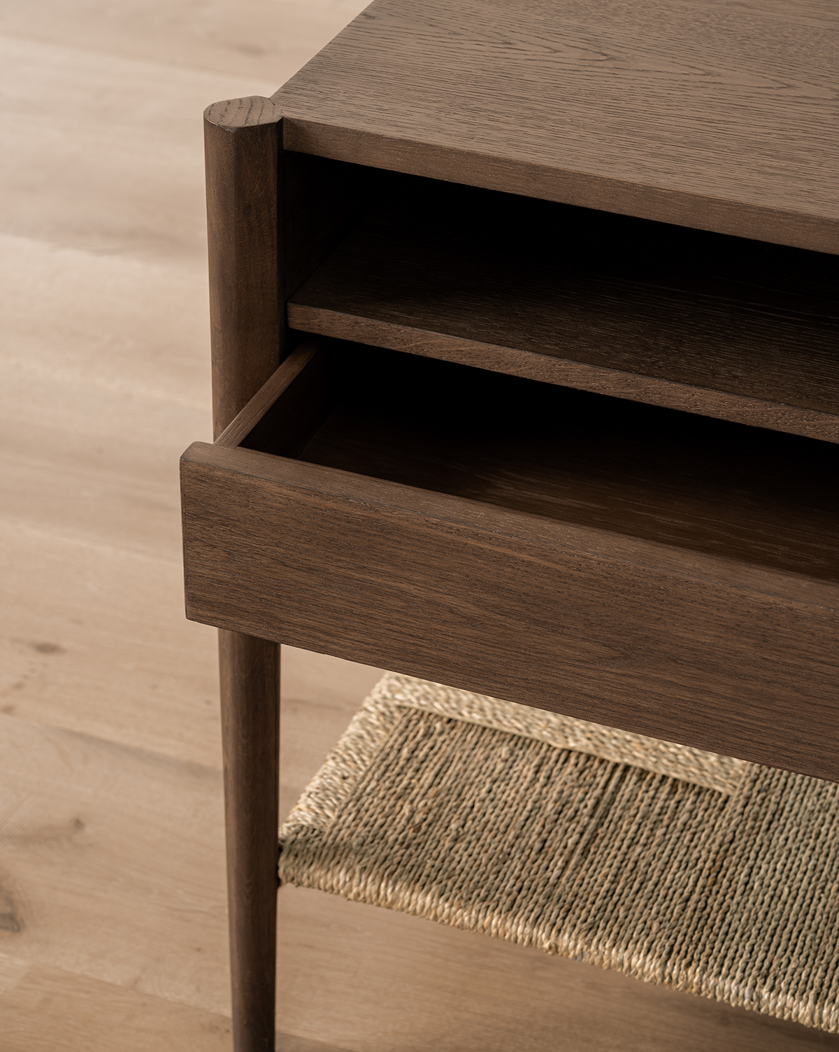 A close-up of the Dana Walnut Oak Nightstand shows its open drawer with brass hardware, shelf above the drawer, and woven rattan lower shelf, all set on light wood flooring, by Vendixs