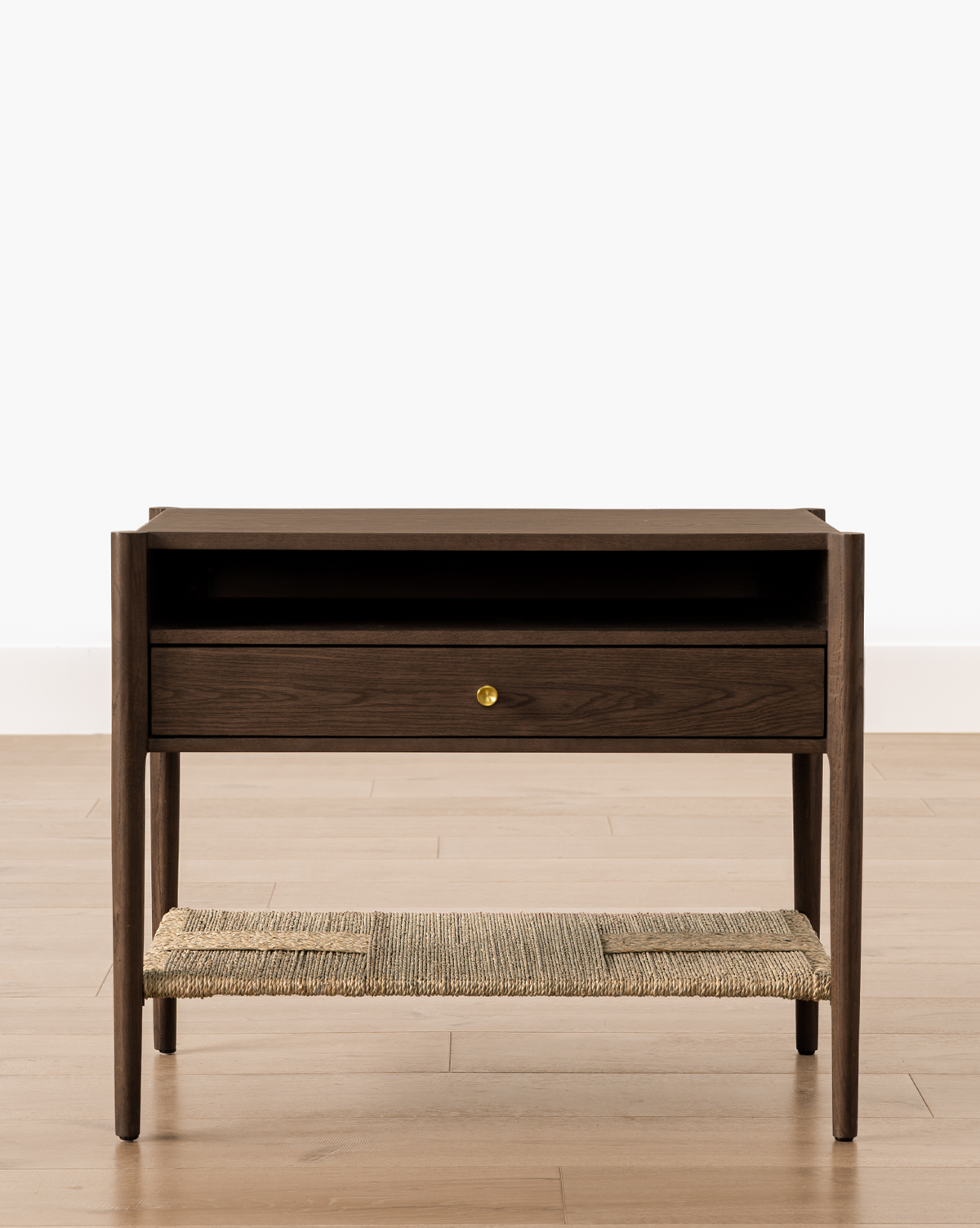 The Dana Walnut Oak Nightstand, featuring brass hardware, a single drawer with a round brass knob, an open shelf above, and a woven rattan lower shelf, stands on a light wooden floor against a white wall, by Vendixs