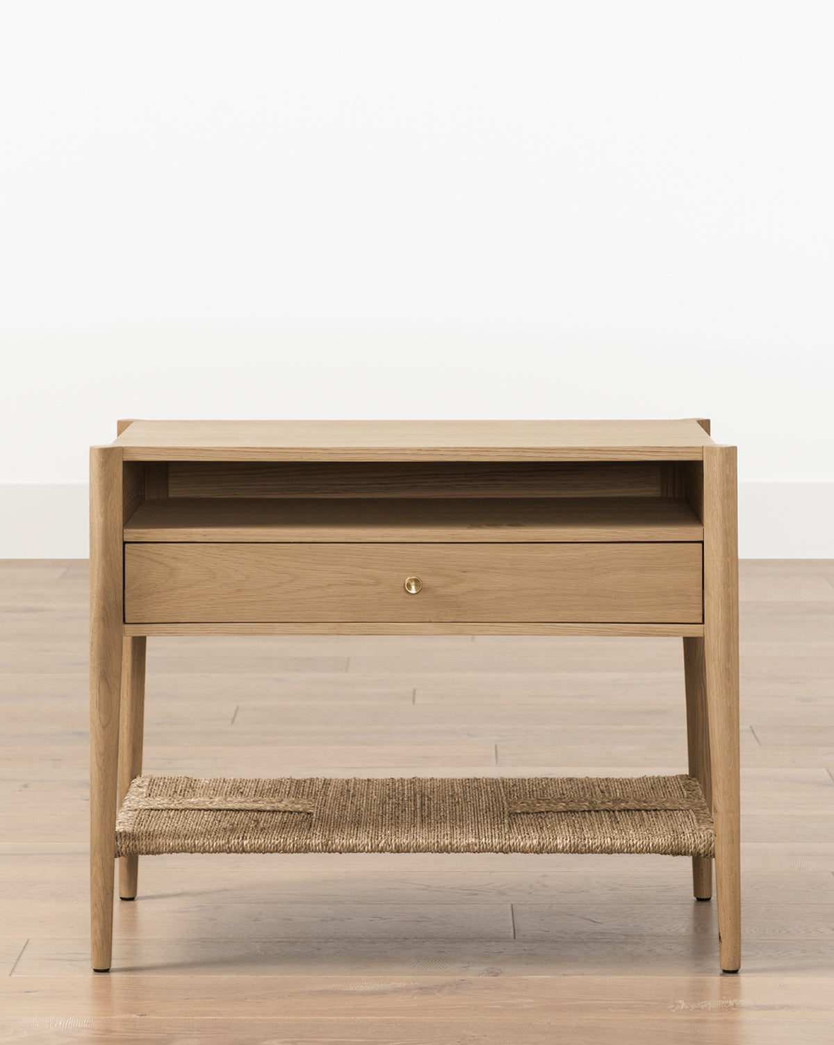 The Dana White Oak Nightstand features a single drawer with brass hardware, an open shelf, and a woven jute lower shelf, by McGee & Co. Its clean, modern lines and four tapered legs stand out against a plain white background.