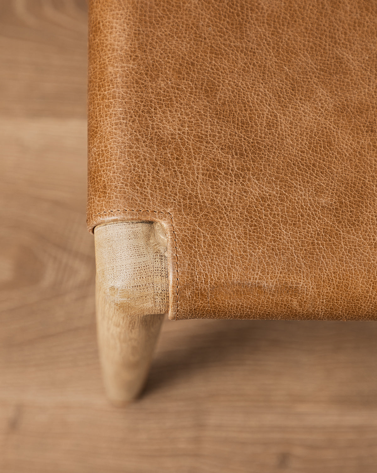 Close-up of the Danson Dining Chair’s wooden leg connected to a light brown, textured leather seat, highlighting its minimalist design on a wood floor, by Vendixs