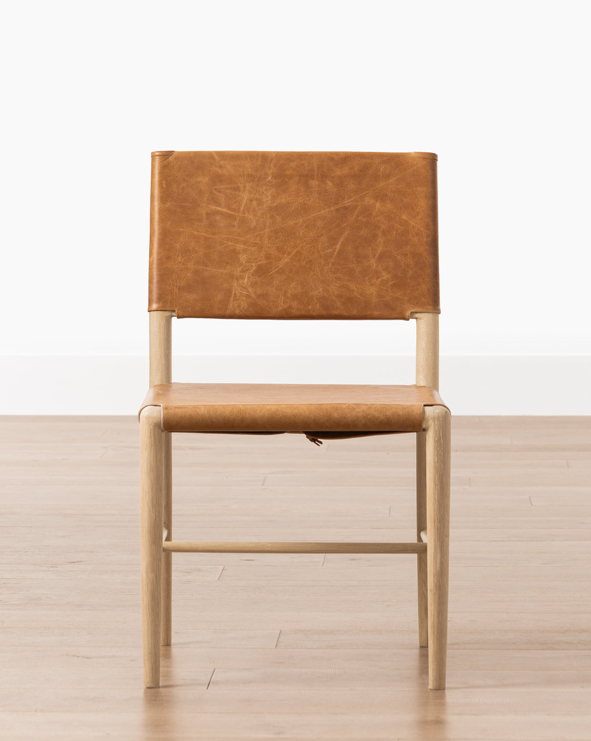 The Danson Dining Chair has a minimalist design with a light wood frame and tan leather seat and backrest, placed on a light wood floor against a plain white wall by McGee & Co.