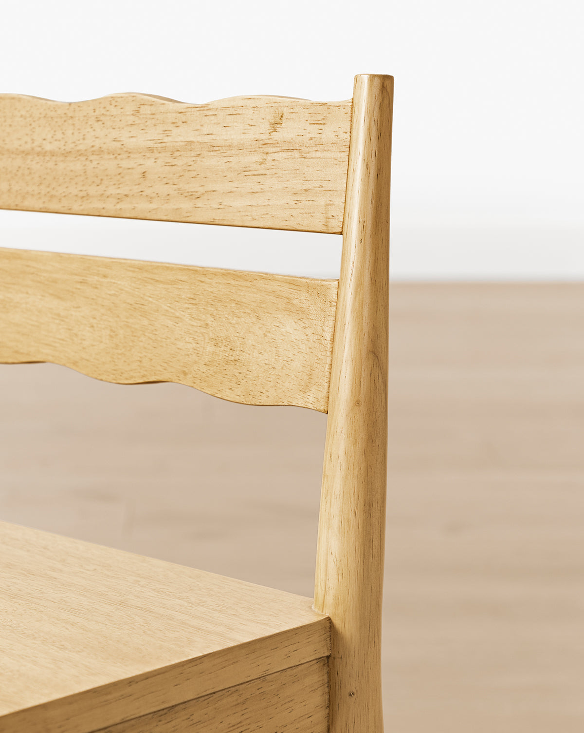 Close-up of the backrest and seat of a Darcy Kids Chair from the Darcy Kids Chairs (Set of 2), featuring a smooth, natural finish and a wavy top slat. The background shows a plain, light-colored floor and wall, by Vendixs