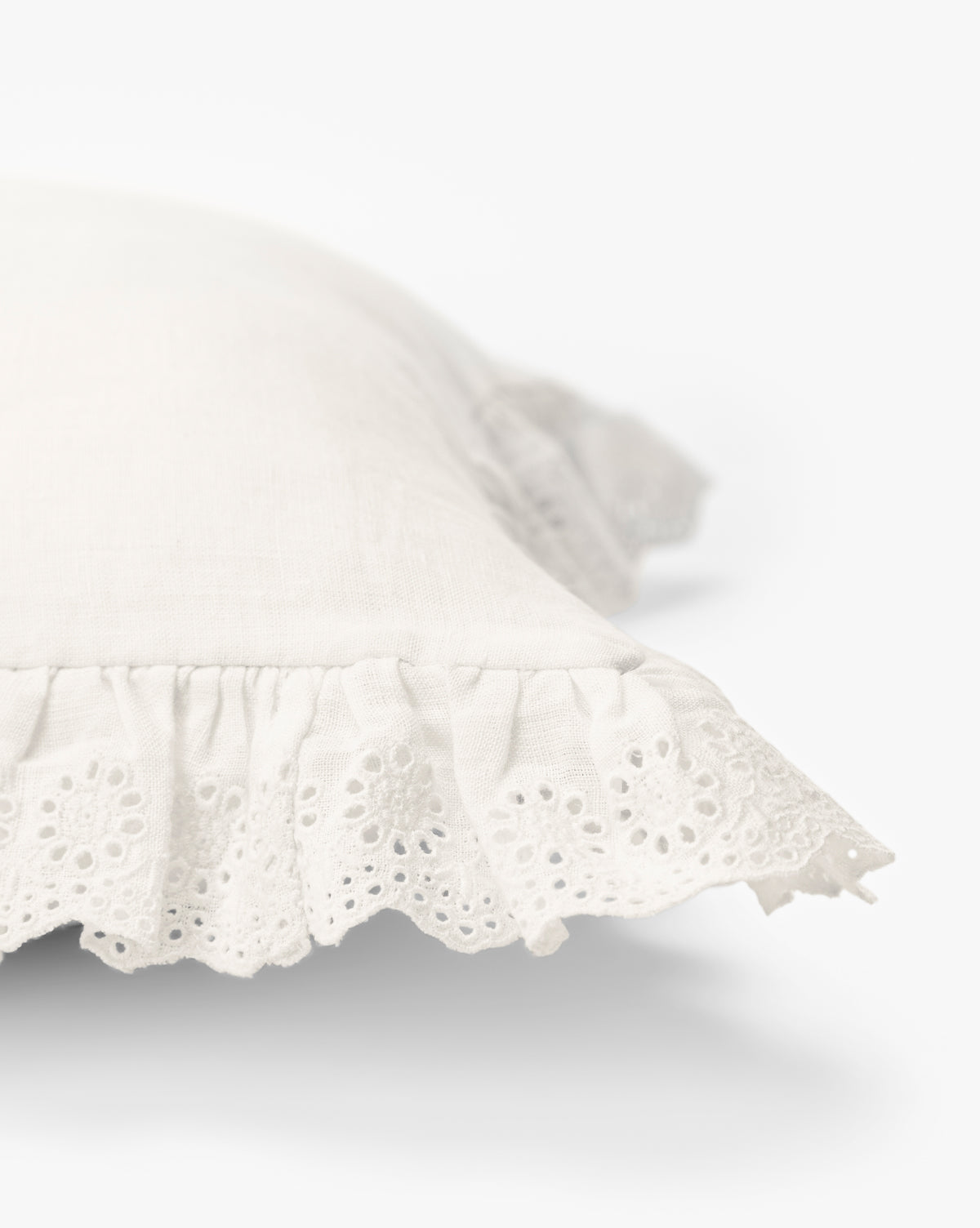 A close-up of the Vendixs x Loeffler Randall Delilah Broderie Trim Pillow Cover shows its white linen, ruffled lace edges, and delicate cut-out embroidery, with a light background that highlights its soft texture and intricate detailing by Vendixs