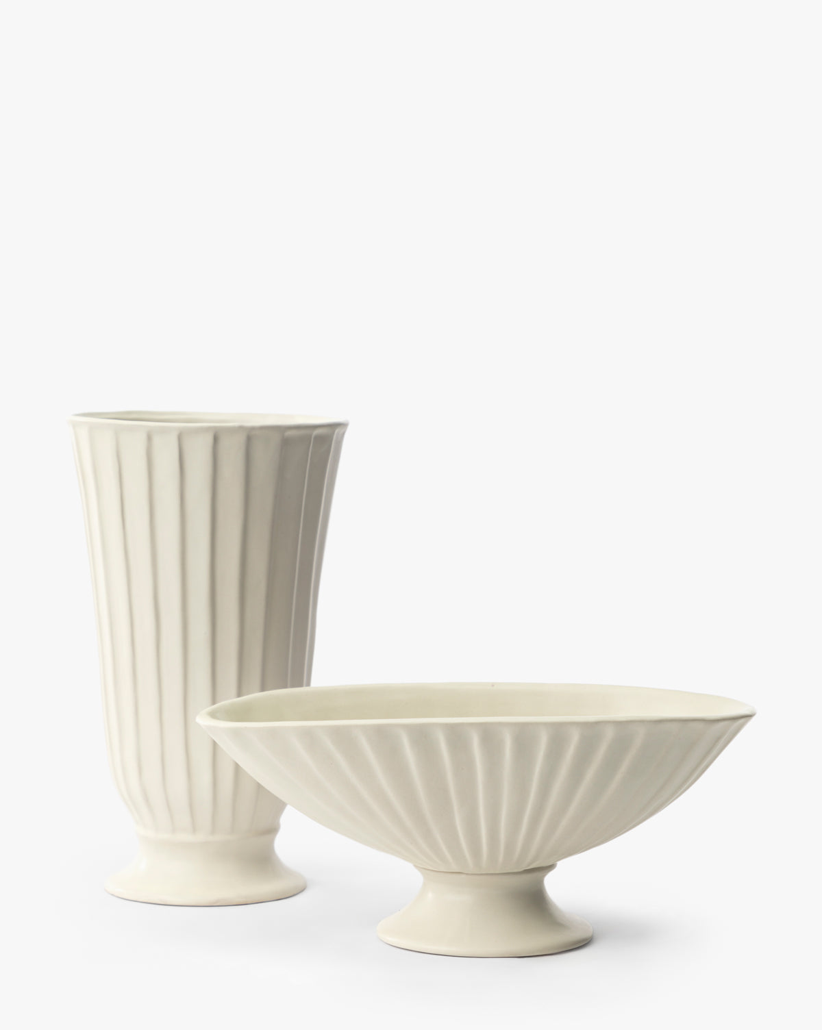 An off-white ceramic set with vertical ridges: a tall, narrow vase stands on the left, and the Delphi Footed Bowl—a wide, fluted vessel with a pedestal base—is placed to the right, both against a plain white background by Vendixs