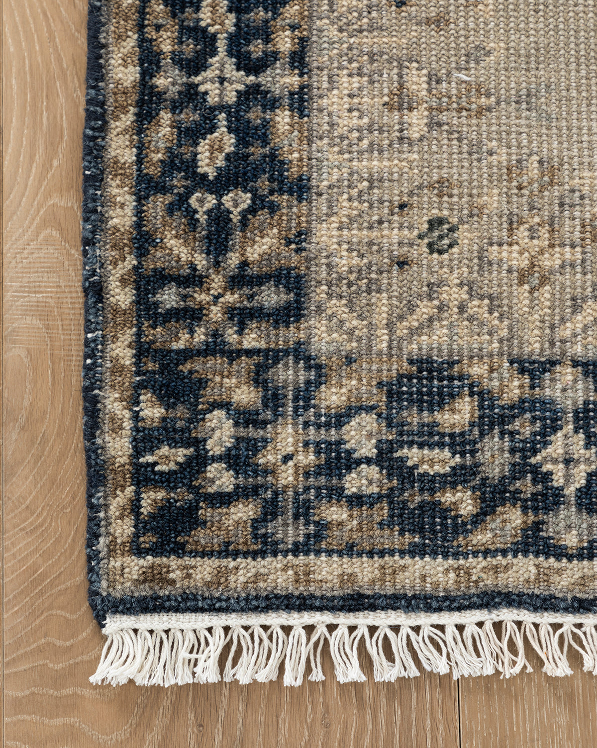 A close-up of the Deveraux Hand-Knotted Wool Rug’s corner shows a dark blue and beige floral border, cream fringe, and muted beige pattern resting on light wood flooring, by McGee & Co.