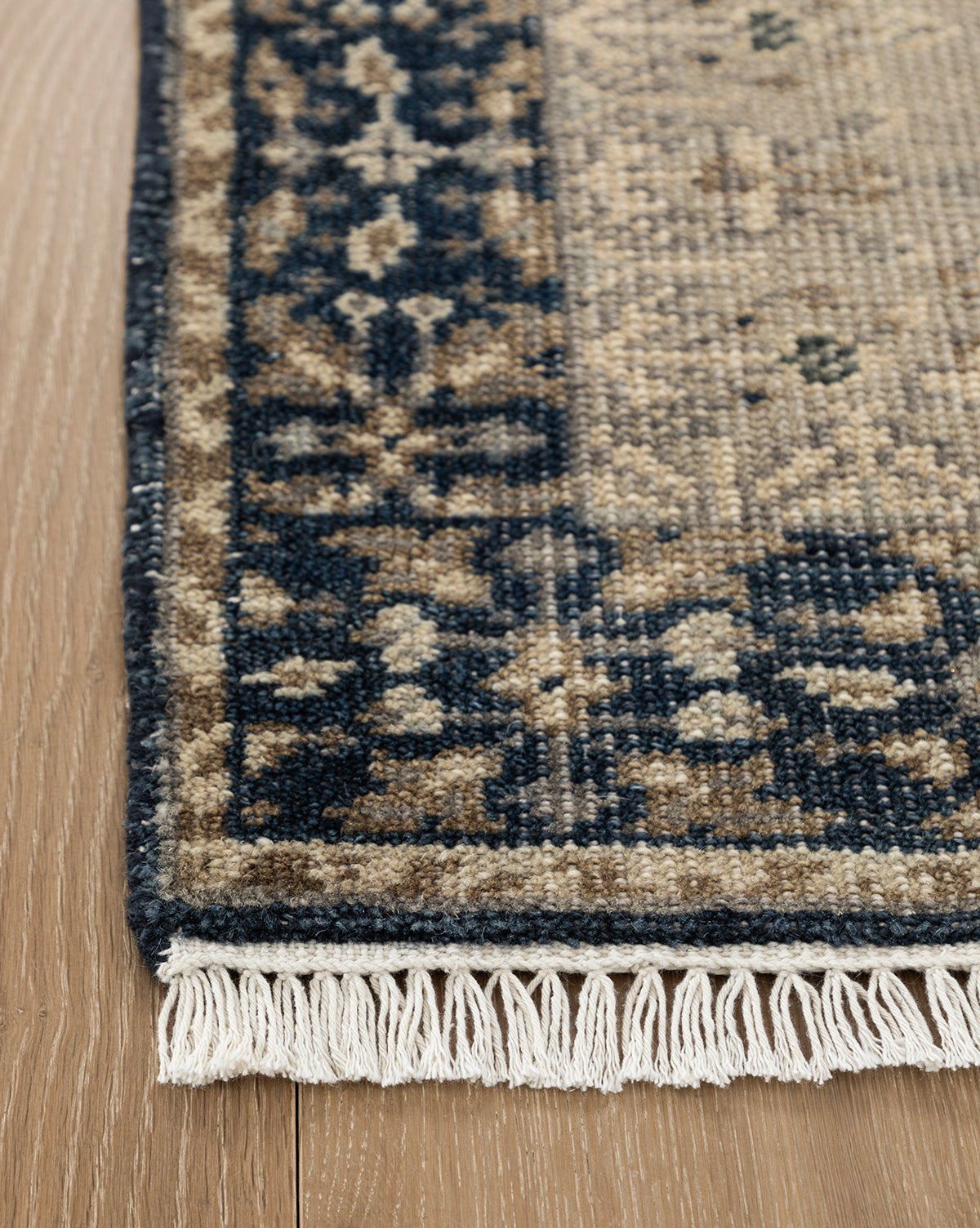 A close-up of the Deveraux Hand-Knotted Wool Rug corner on a wooden floor, showcasing beige, navy, and brown hues with fringe along the edge, by McGee & Co.