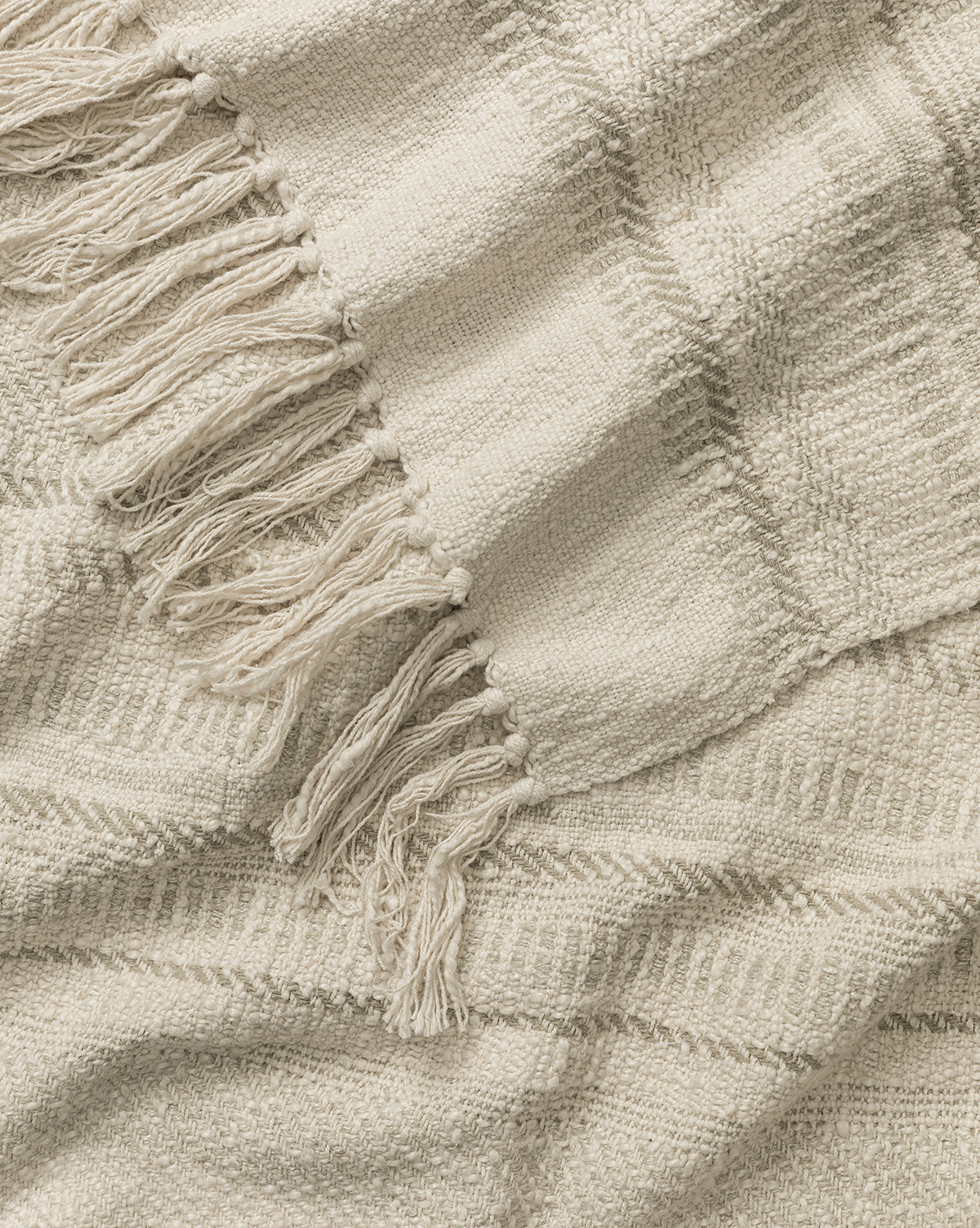 A close-up of the Donahue Striped Throw by ILA HOME FASHIONS, a cream cotton-blend blanket with fringed edges and subtle stripes. This soft, cozy throw adds warmth and texture to any space—Studio Vendixs