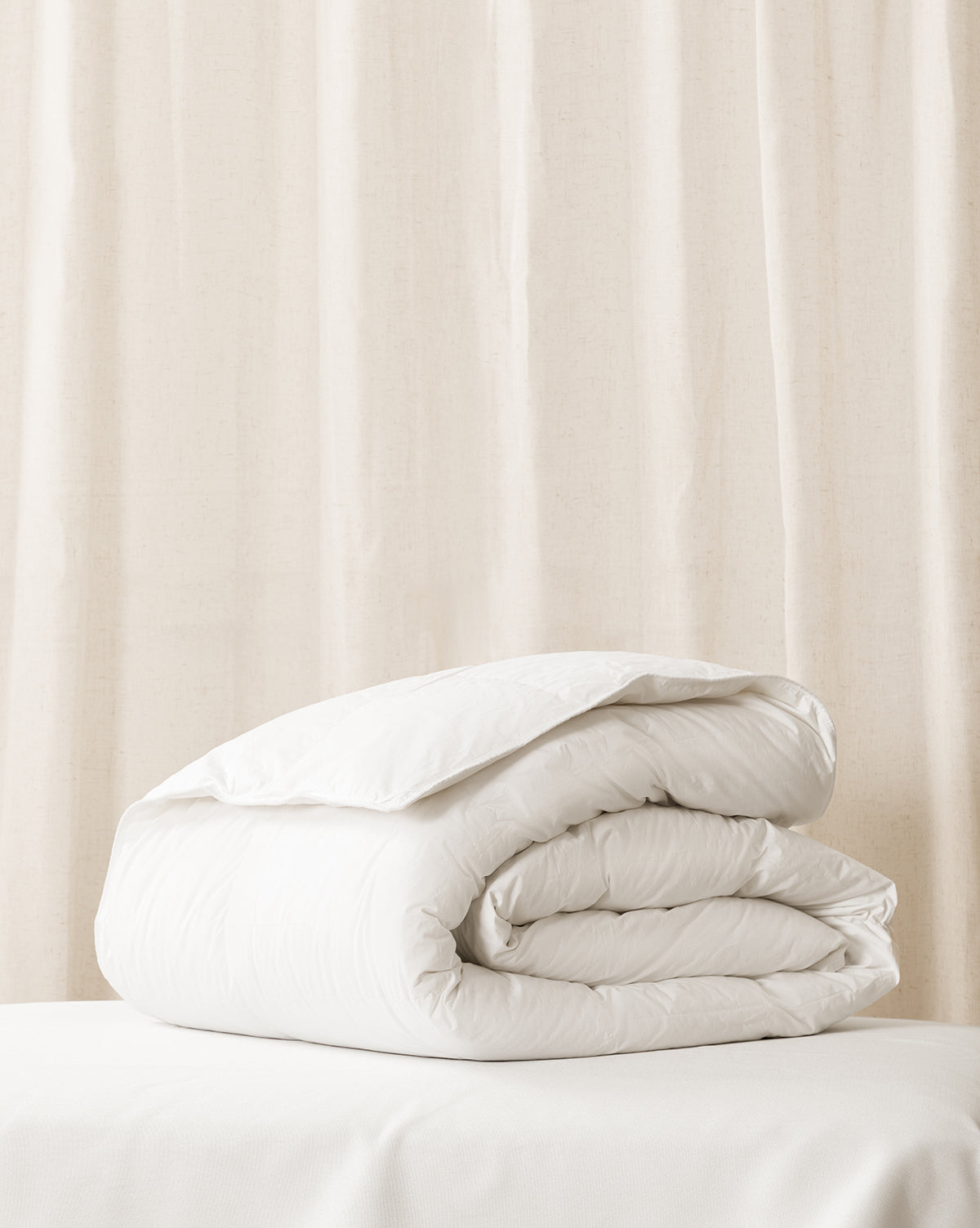 A folded white down duvet insert on a bed with a beige curtain in the background from Vendixs