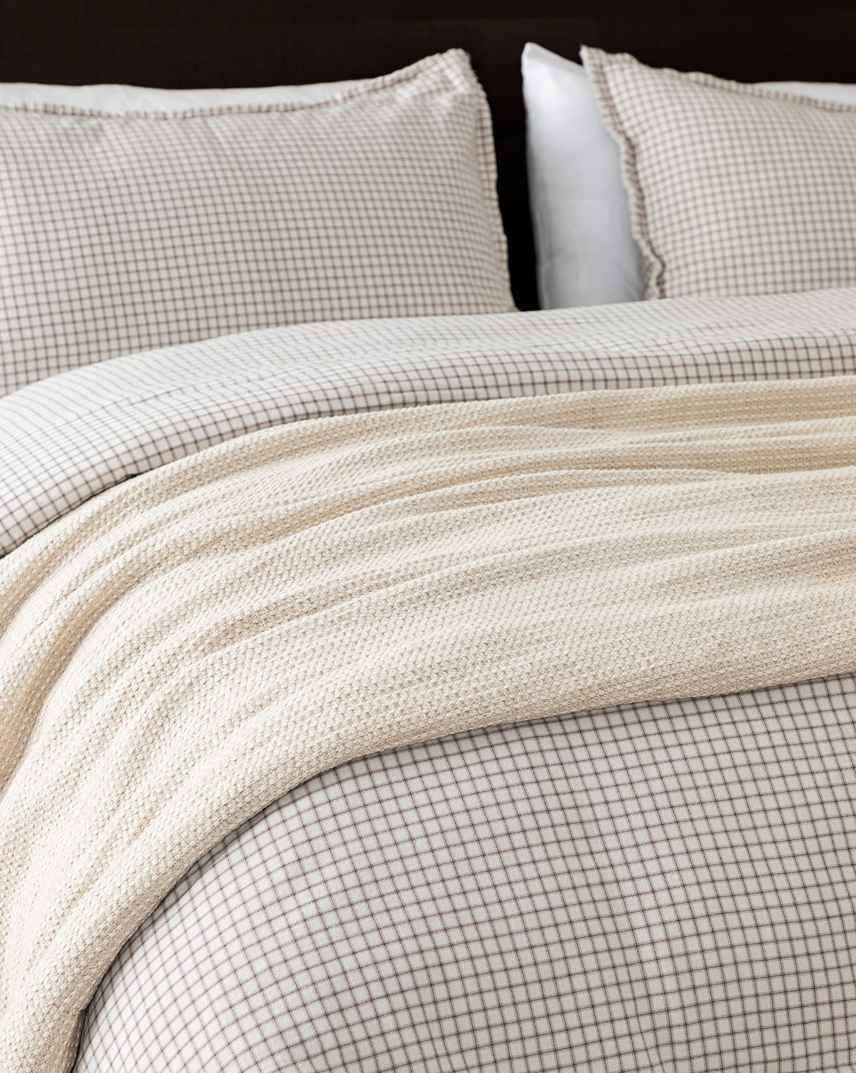 A neatly made bed with a beige and white small checkered duvet and matching pillows is topped with the Penedo Duffy Oversized Throw, a cream-colored knit blanket in neutral tones draped across the foot of the bed, Vendixs