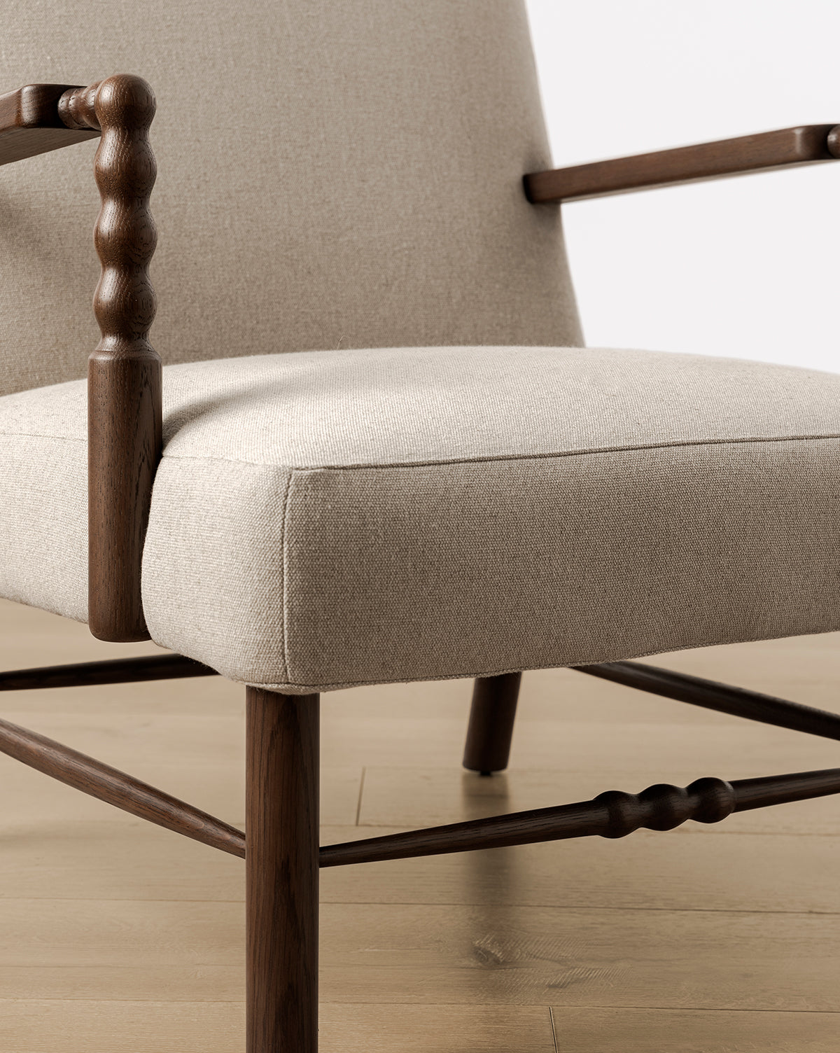 Close-up of the Earnshaw Lounge Chair, showcasing neutral upholstery and wooden armrests and legs in English oak with turned wood details, set on a light wood floor against a neutral backdrop, by Vendixs