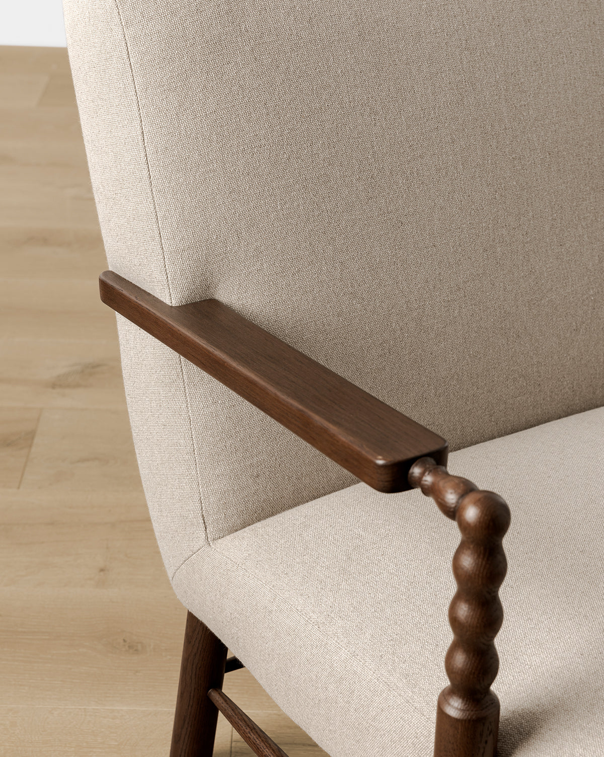 A close-up of the Earnshaw Lounge Chair shows its neutral upholstery and dark English oak armrest with spindle-style support, set on a light wood floor, by Vendixs