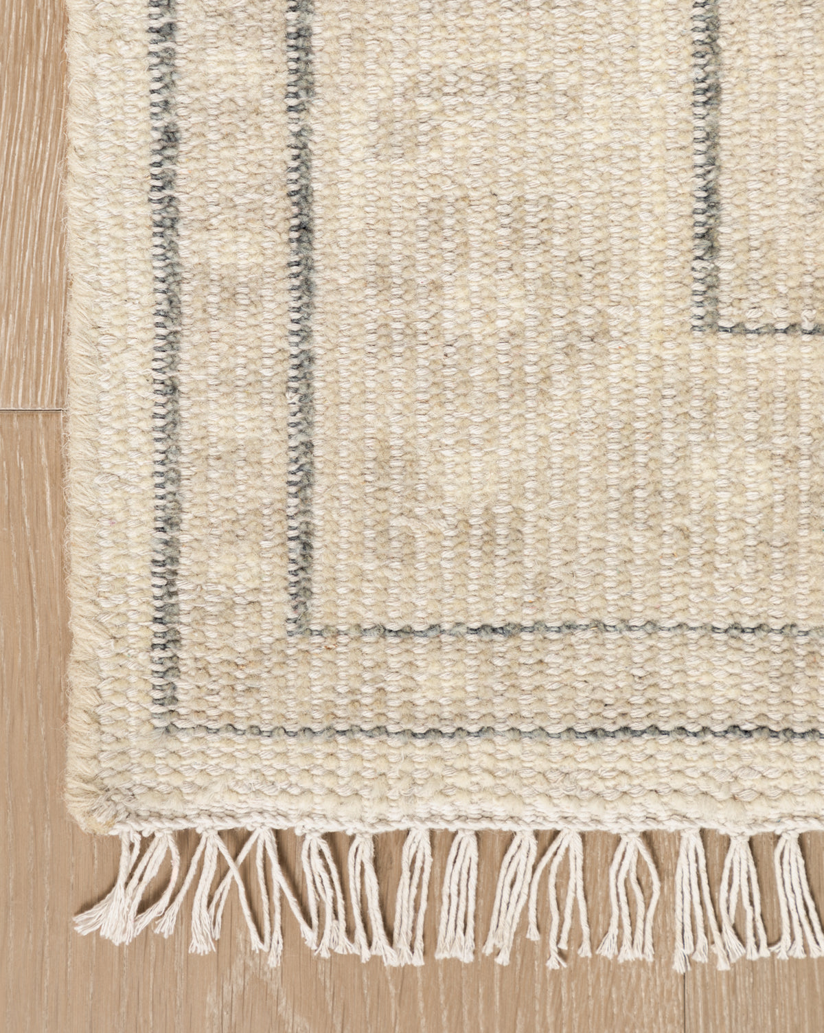 A close-up of the Elin Hand-Knotted Wool Rug highlights its simple gray border, artisan-crafted wool texture, and fringe details, beautifully displayed on a light wooden floor, by Vendixs