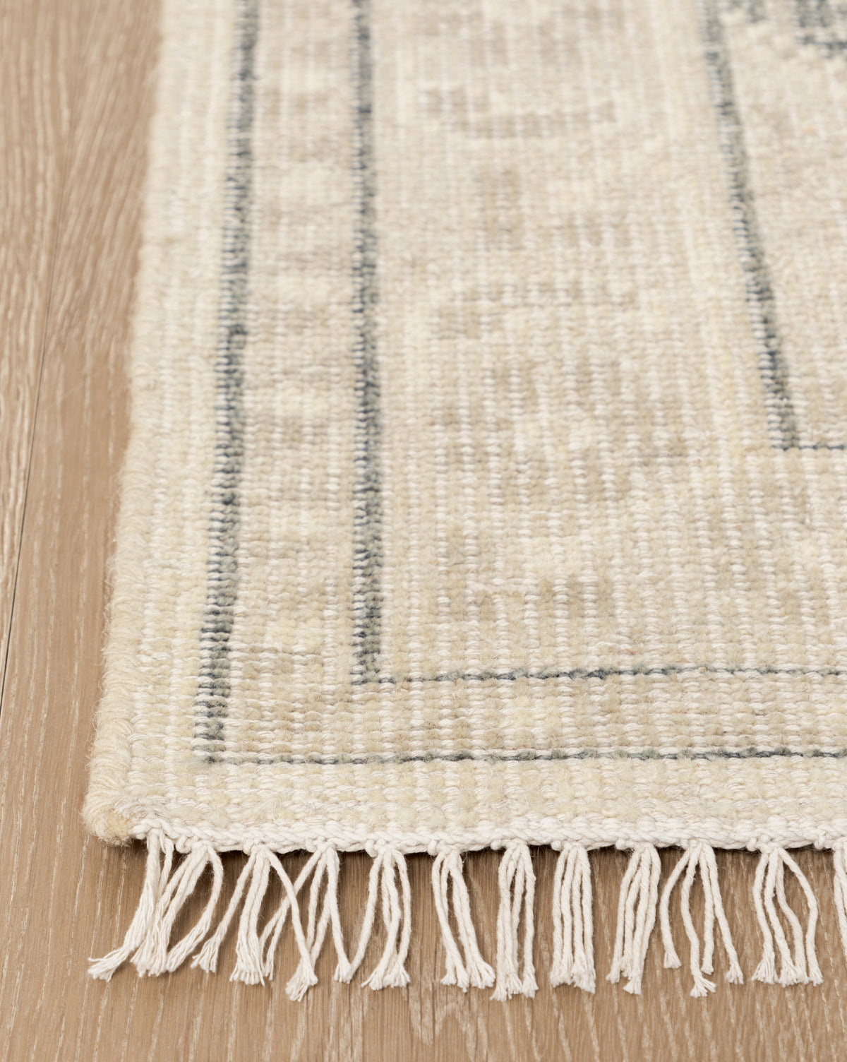 A close-up of the Elin Hand-Knotted Wool Rug, showing its dark border lines and white fringes with artisan details, placed on a light wood floor, by Vendixs