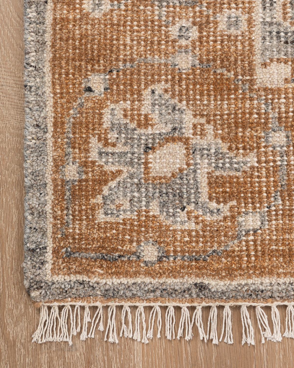 A close-up of the Elison Hand-Knotted Wool Rug corner displays geometric and floral patterns in brown, gray, and cream with short fringe on wood flooring, emphasizing its artisan craftsmanship, by Vendixs