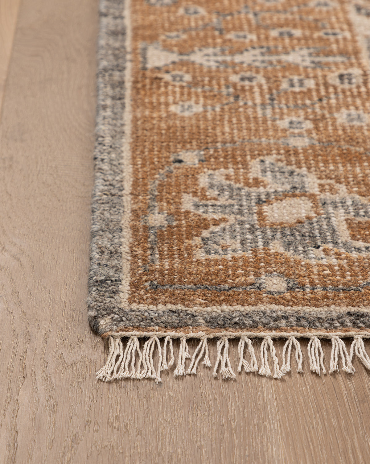 Close-up of the Elison Hand-Knotted Wool Rug’s corner with fringe on light wood flooring. The rug displays earthy brown and gray tones, a geometric pattern, and a decorative gray border, by McGee & Co.