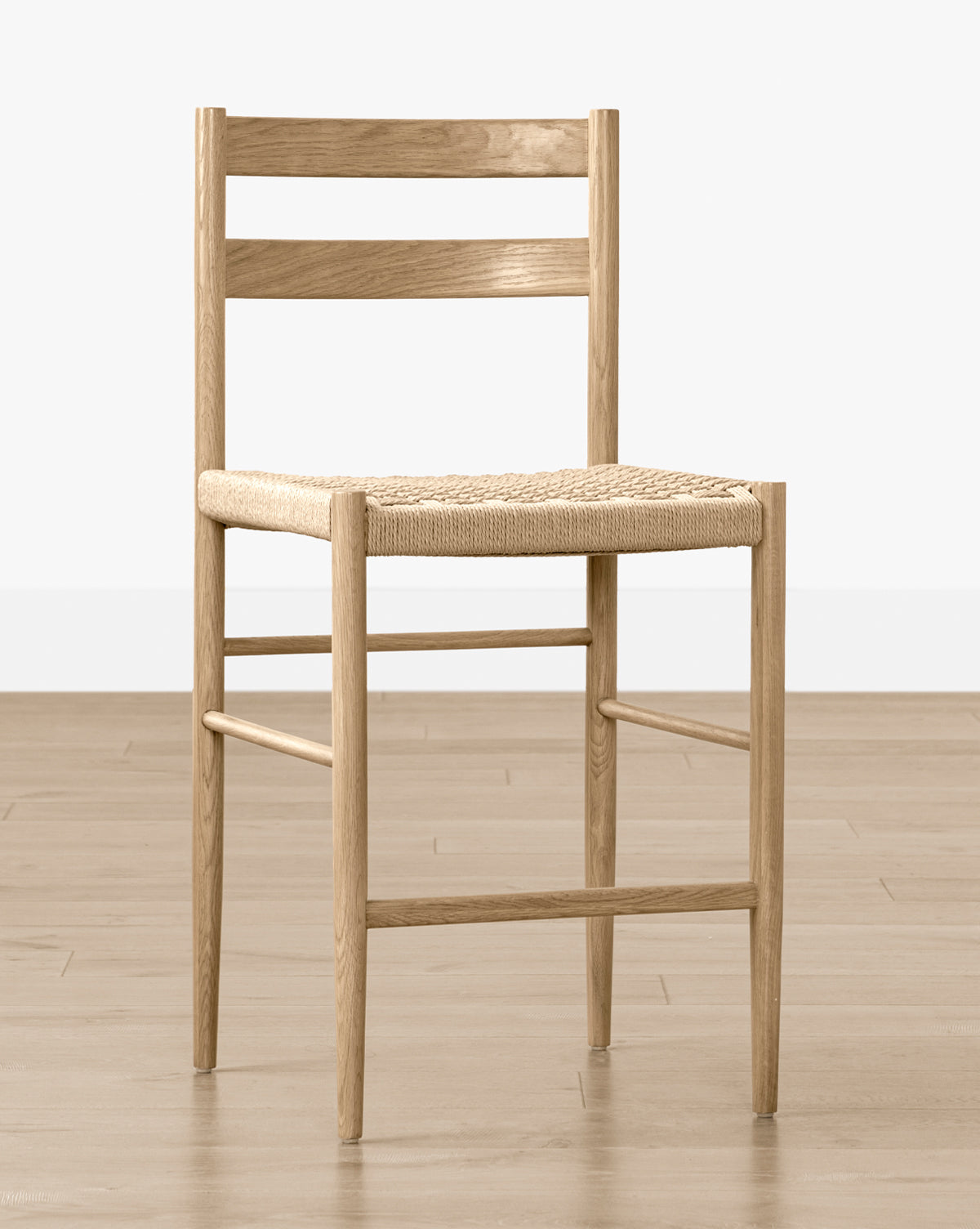 The Eloise Woven Counter Stool features a light oak frame, straight legs, horizontal backrest slats, and a woven seat in classic Danish design, shown on a wooden floor against a plain white wall by McGee & Co.