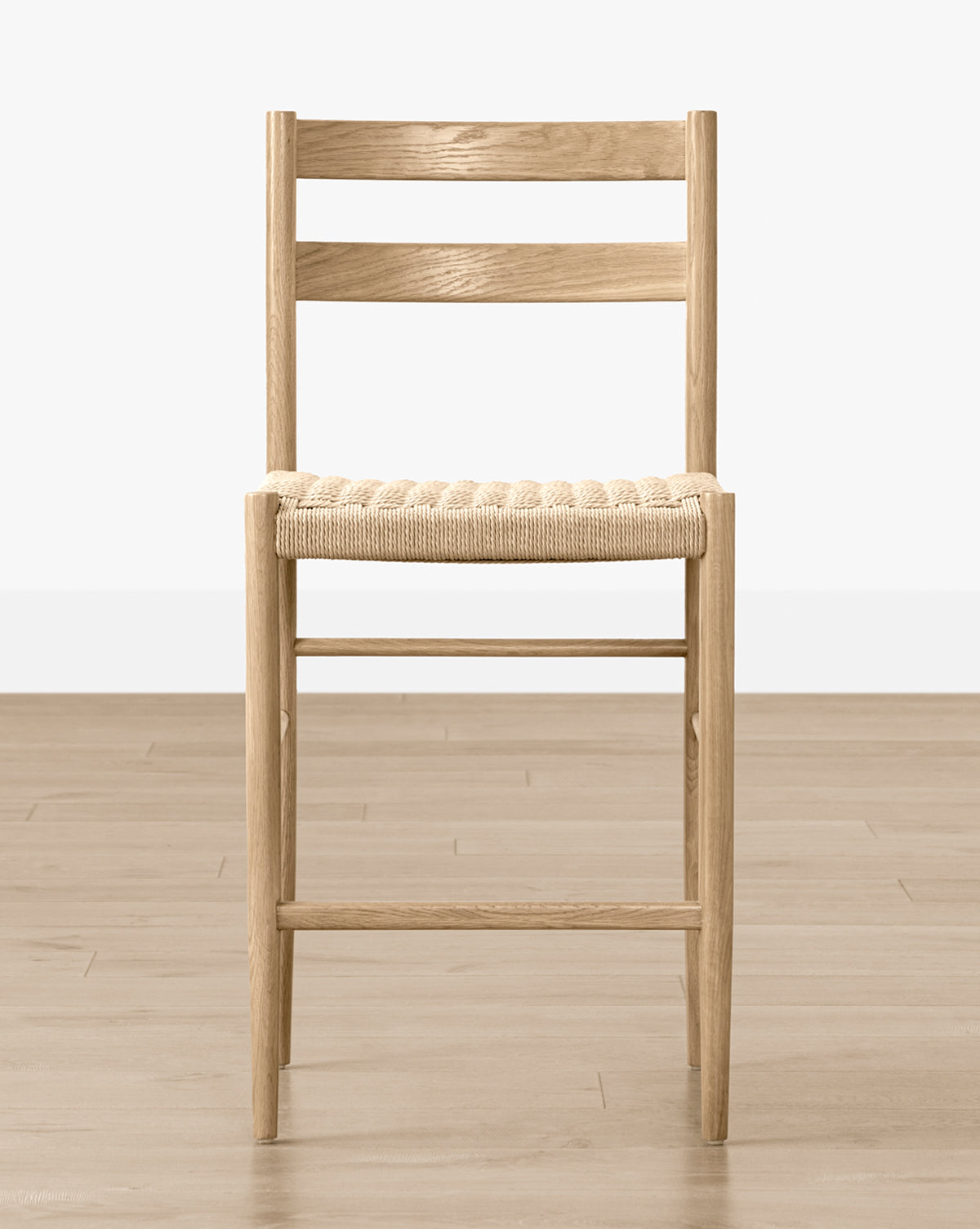 The Eloise Woven Counter Stool, featuring a wooden frame and woven seat with simple straight lines, sits on a light wood floor against a white wall, showcasing classic Danish design, by McGee & Co.