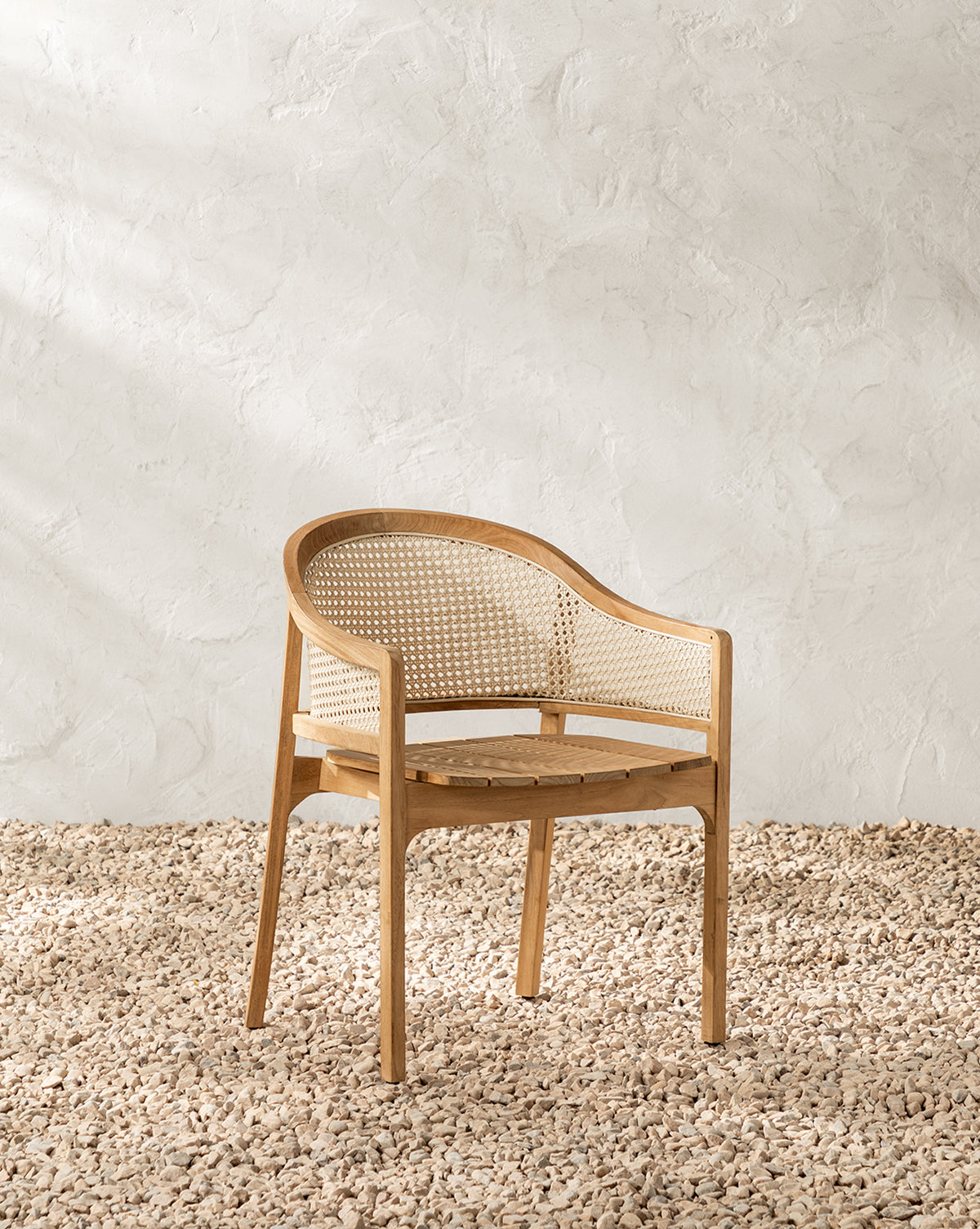 The Elowyn Outdoor Dining Chair, featuring a curved backrest and cane woven seat, sits on light-colored pebbles with a textured white wall as the backdrop, by Vendixs