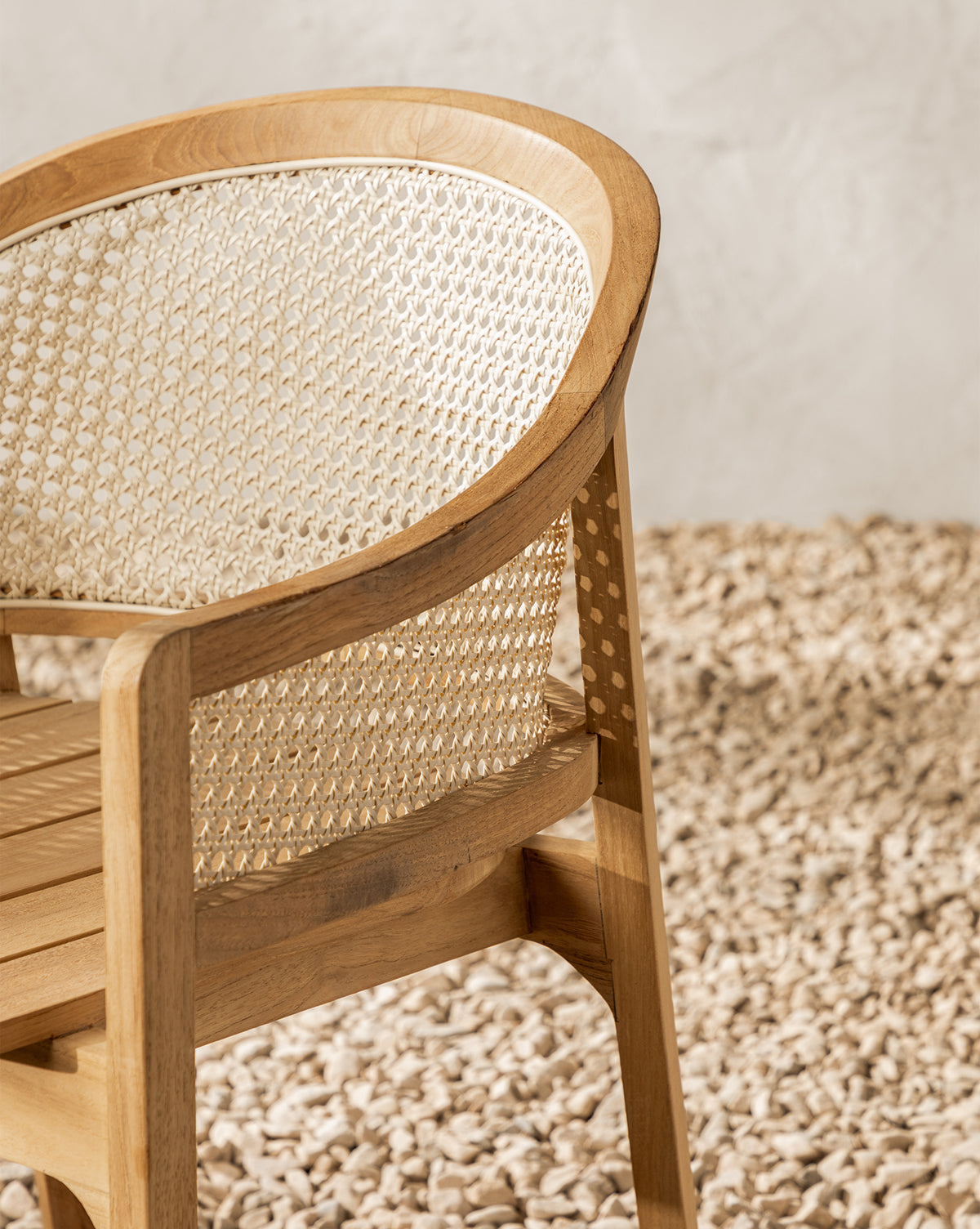 A close-up of the Elowyn Outdoor Dining Chair shows its curved cane backrest and teak finish, set on small, light-colored stones with a subtly patterned light wall in the background, by Vendixs