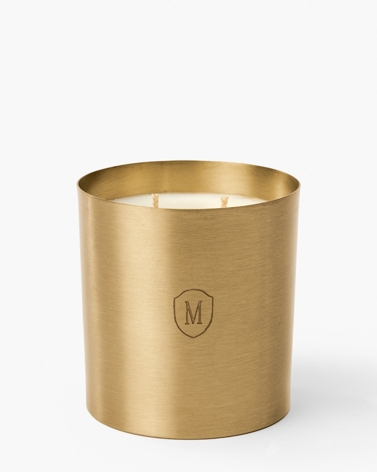 The Embossed Brass Candle features a gold artisan holder with a white candle inside, two unlit wicks, and an engraved shield with the letter M on the front for a refined display, by Vendixs
