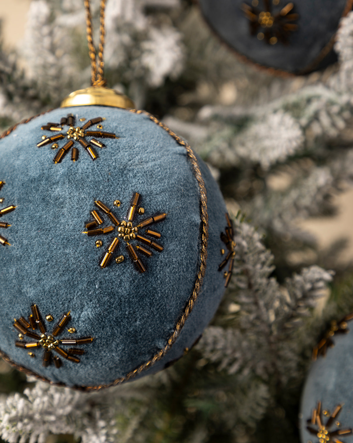 A 4" Embroidered Velvet Bauble from a set of 3, featuring blue velvet with gold beaded snowflakes, brings vintage elegance to a snow-dusted Christmas tree, by Vendixs