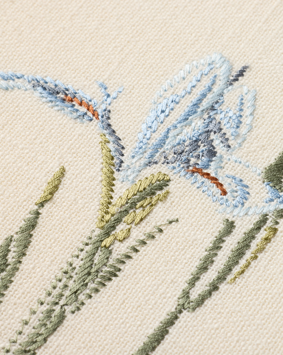 Close-up of the Embroidered Blue Iris features delicate cotton embroidery on beige fabric, with blue iris flowers, green stems and leaves, and subtle orange accents. The textured threads enhance this botanical wall art, by Vendixs
