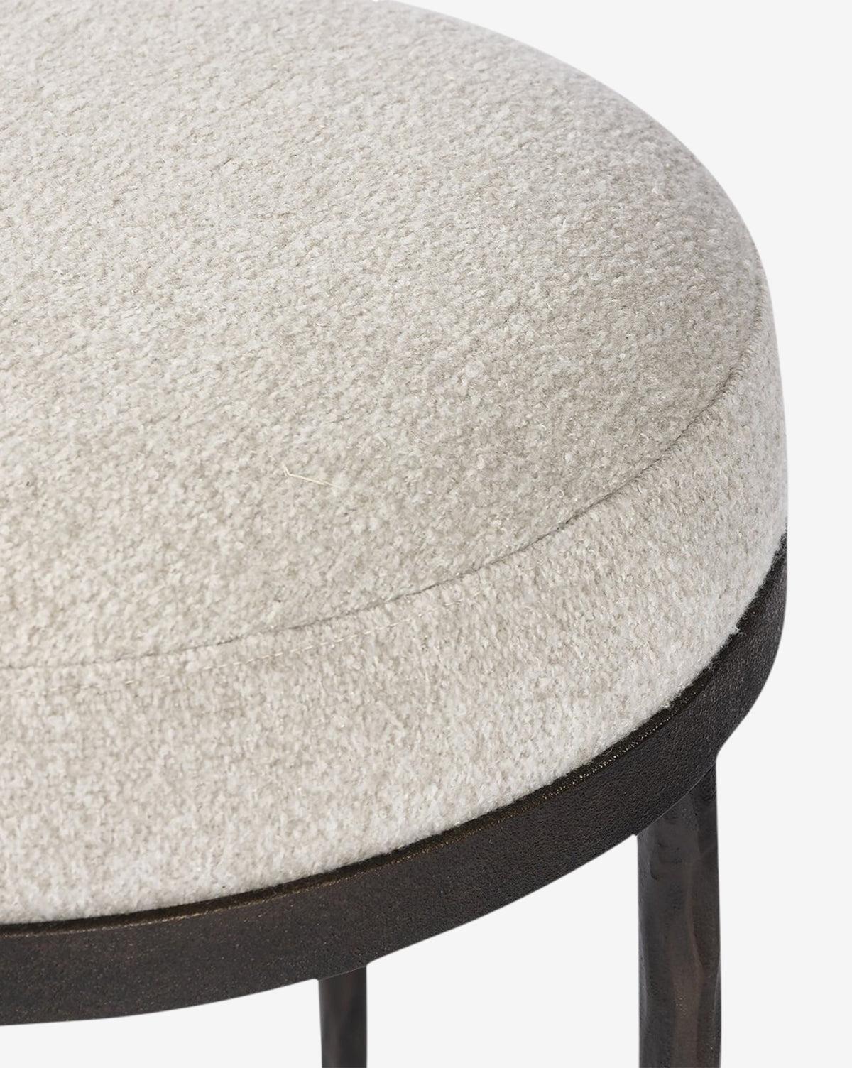 Close-up of the Emmett Stool by Four Hands—a round, upholstered cocktail ottoman featuring textured off-white fabric and a dark hammered bronze base with slim legs, making it a stylish accent for any room. Vendixs