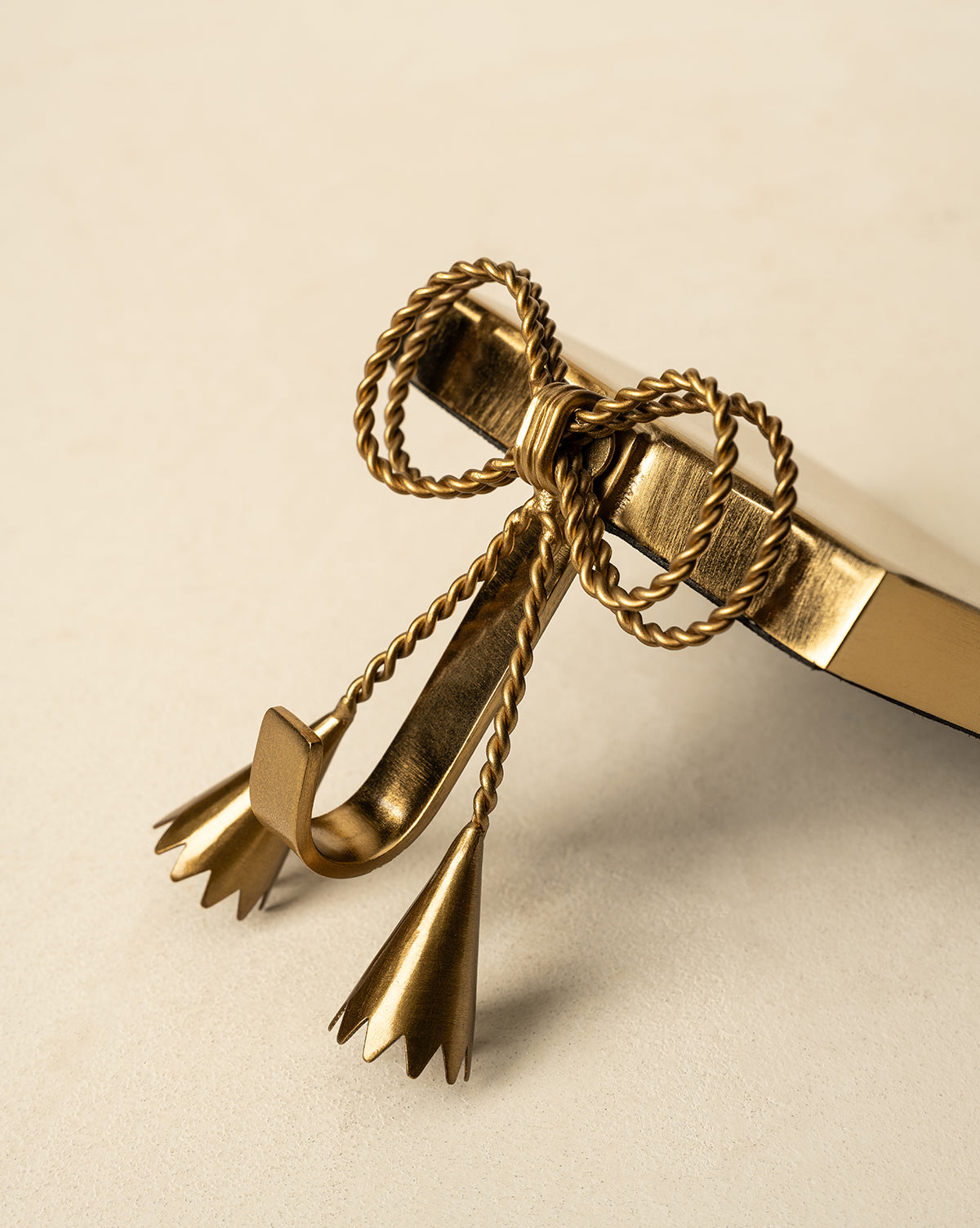 A gold metal book hook featuring a twisted rope bow and tassel design, inspired by the Erika Stocking Holder, rests on the corner of a closed book against a neutral background, by Vendixs