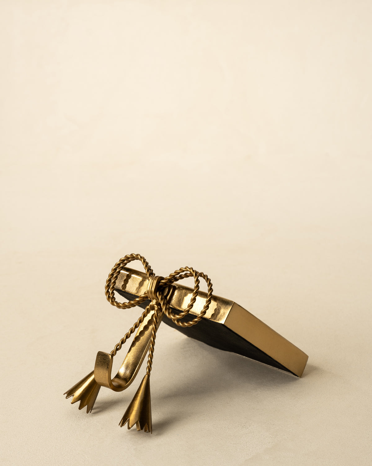 The Erika Stocking Holder, featuring an antique brass finish and decorative rope bow with tassels, is displayed on a neutral beige surface against a light background, by Vendixs