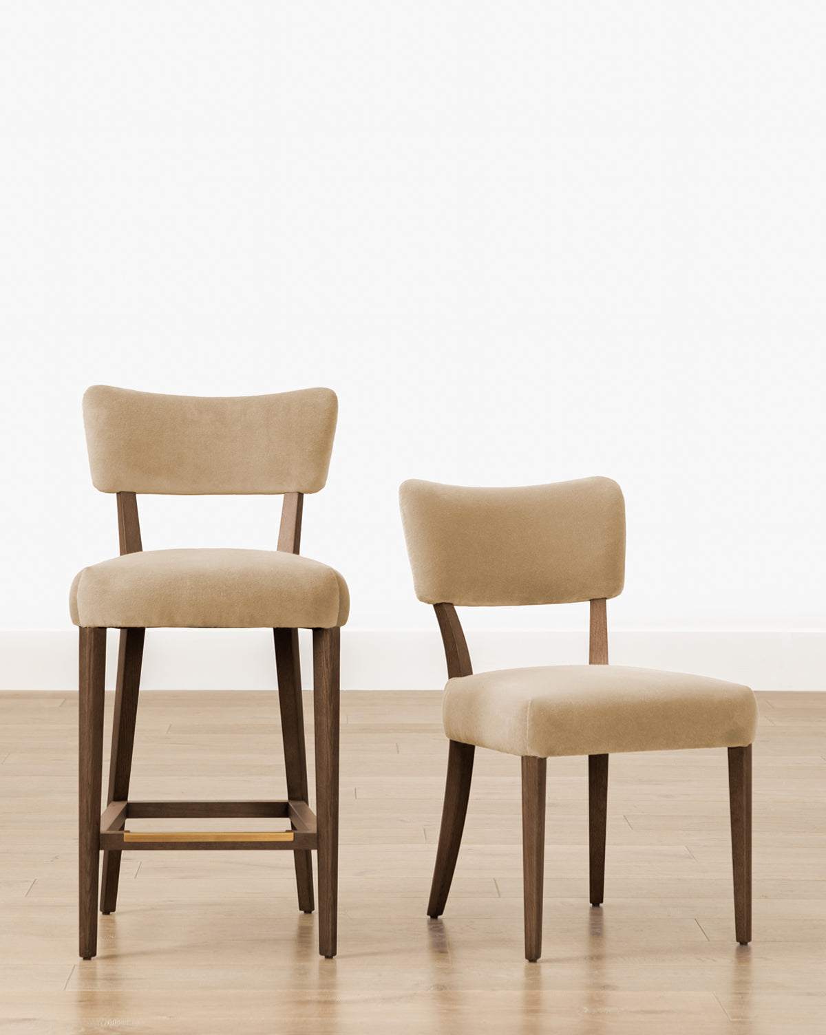 Two beige upholstered chairs with wooden legs sit on a light wood floor: the Makerspalm Etienne Stool (Ready to Ship), a tall, vintage-inspired kitchen stool, and a standard-height dining chair. Both feature curved backs and minimalist design from Vendixs
