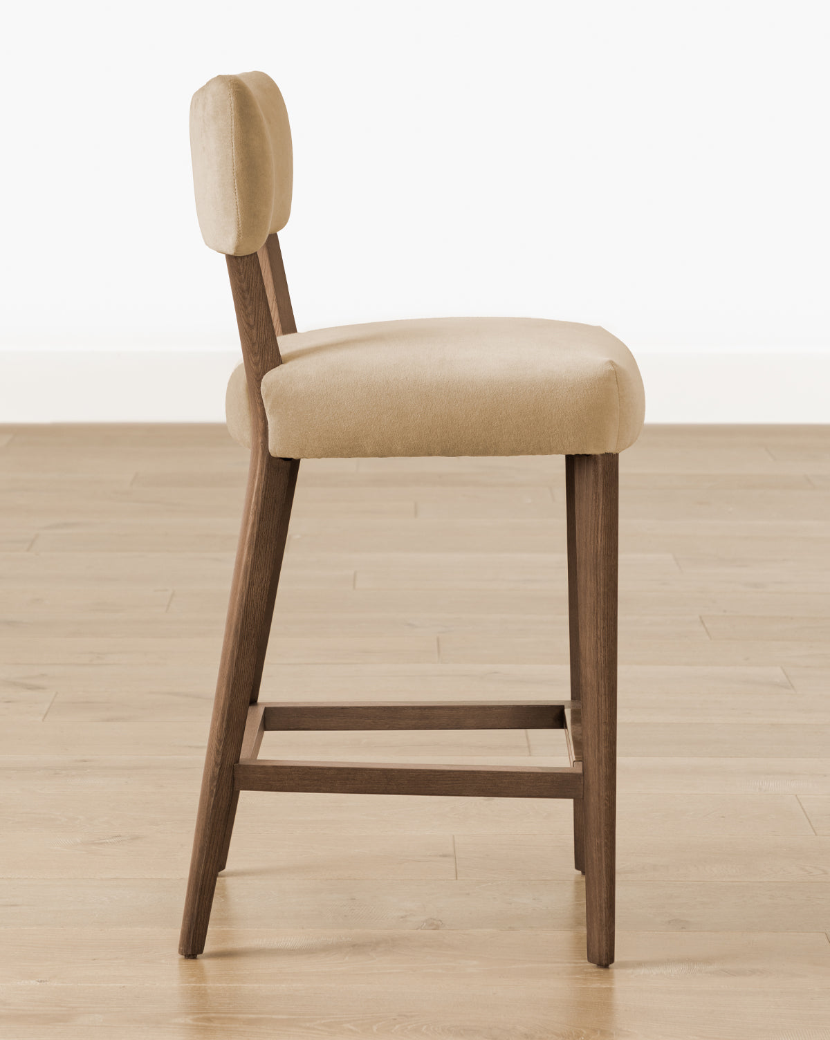 Certainly! Here’s your blended description with the exact string at the end: Side view of the Makerspalm Etienne Stool (Ready to Ship), featuring a light beige cushioned seat and wooden angled legs, displayed on a light wood floor with a plain white wall background. Vendixs