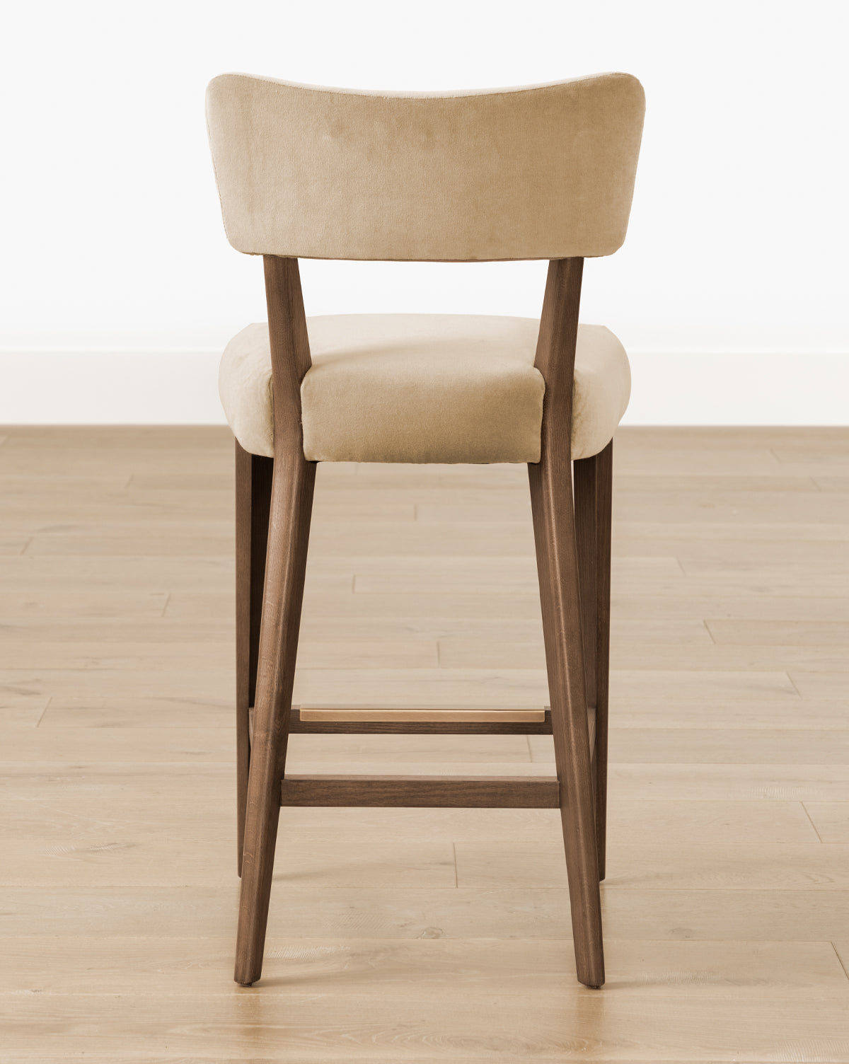 The Etienne Stool (Ready to Ship) by Makerspalm features beige upholstery, a curved backrest, and wooden legs, shown from the back on a light wood floor with a white wall background. Vendixs