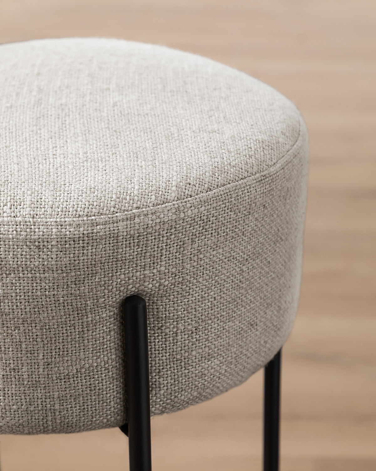 Close-up of the Makerspalm Evander Counter Stool, featuring industrial design with light gray textured fabric and black metal legs, set against a blurred wooden floor—ideal for stylish modern kitchen seating. Vendixs