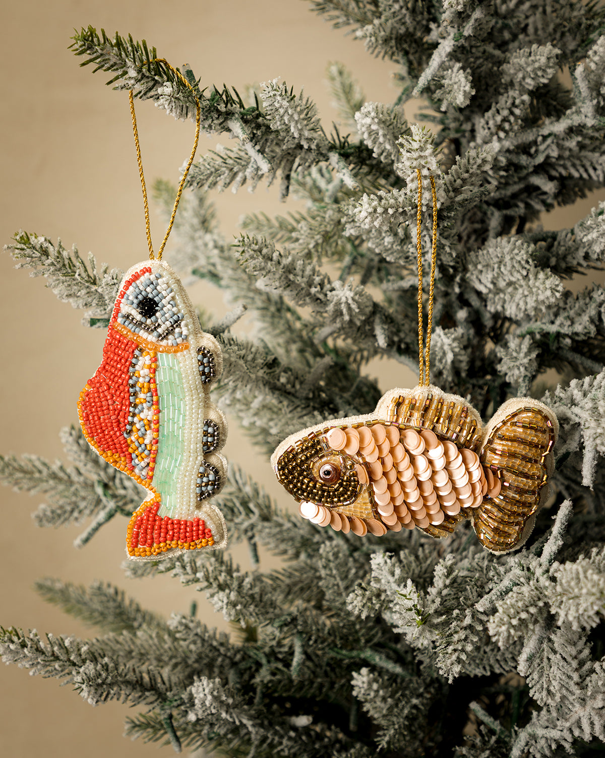 The Fabric Sequin Fish Ornaments (Set of 2), featuring one multicolored with hand-applied beads and one gold and cream, add festive charm to a snow-frosted artificial Christmas tree—perfect for holiday decorating, by Vendixs
