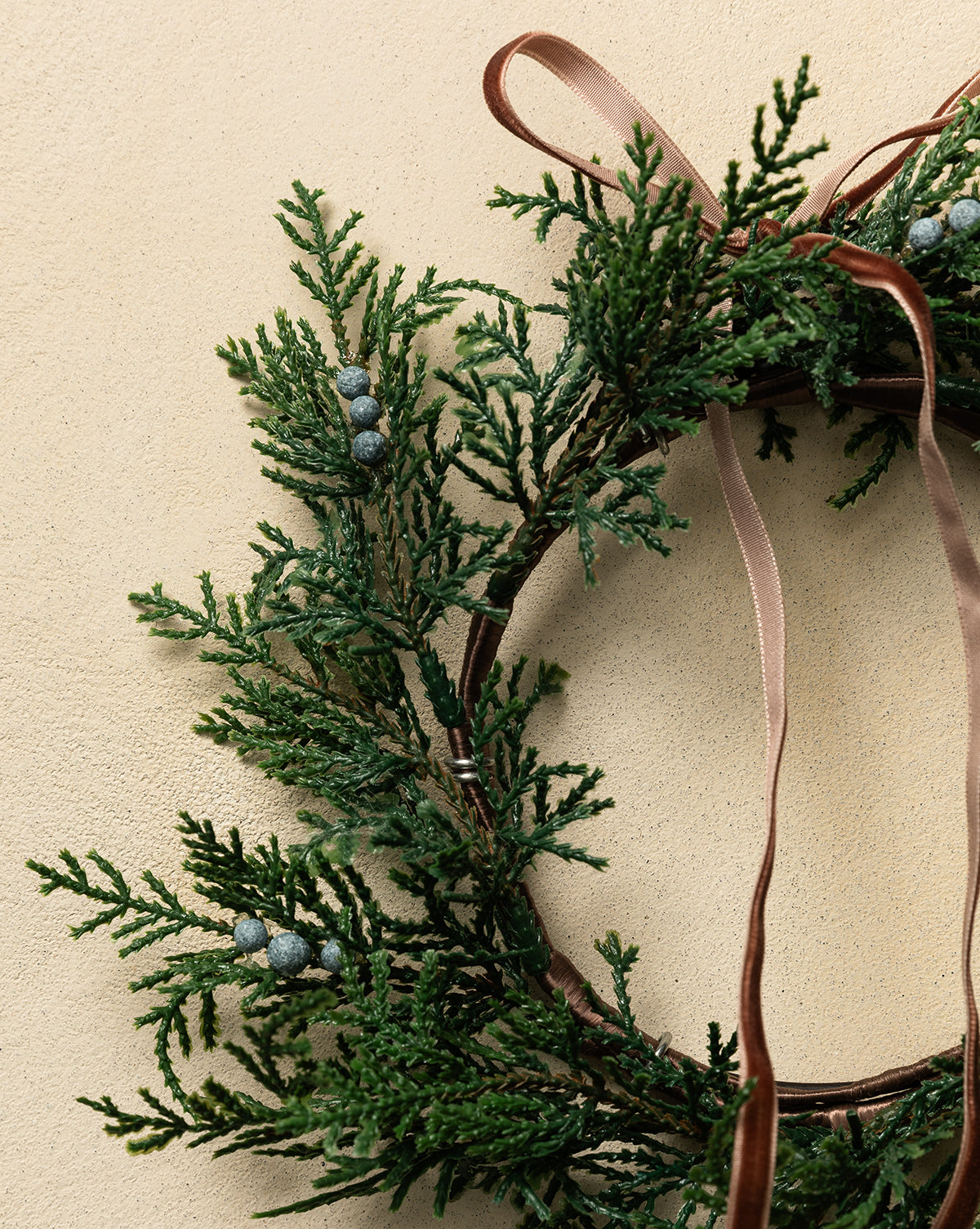 A close-up of the 8" Blueberry & Juniper Faux Wreath shows faux greenery, blue berries, and a brown ribbon on a light beige textured background—ideal for stylish holiday décor, by Vendixs