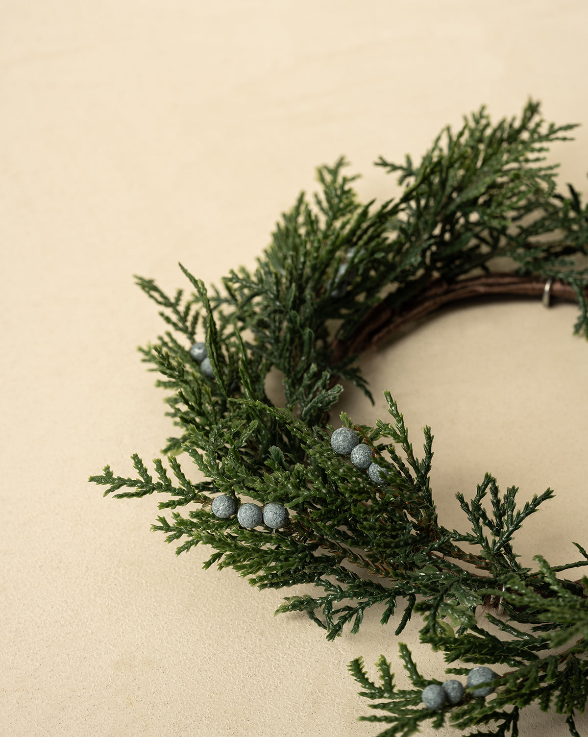A partial view of the 8" Blueberry & Juniper Faux Wreath, featuring green pine branches and gray berries on a light beige background—an ideal faux greenery accent for elegant holiday decor, by Vendixs