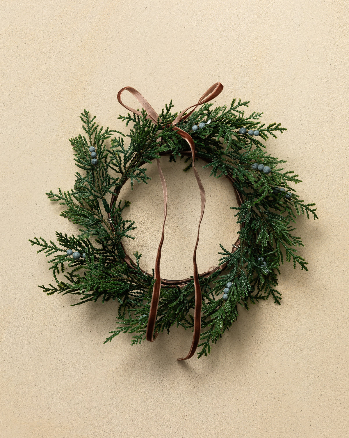 The 8" Blueberry & Juniper Faux Wreath, with lush faux greenery, blueberries, and a brown ribbon bow, adds a charming holiday touch as it hangs on a light beige wall, by Vendixs