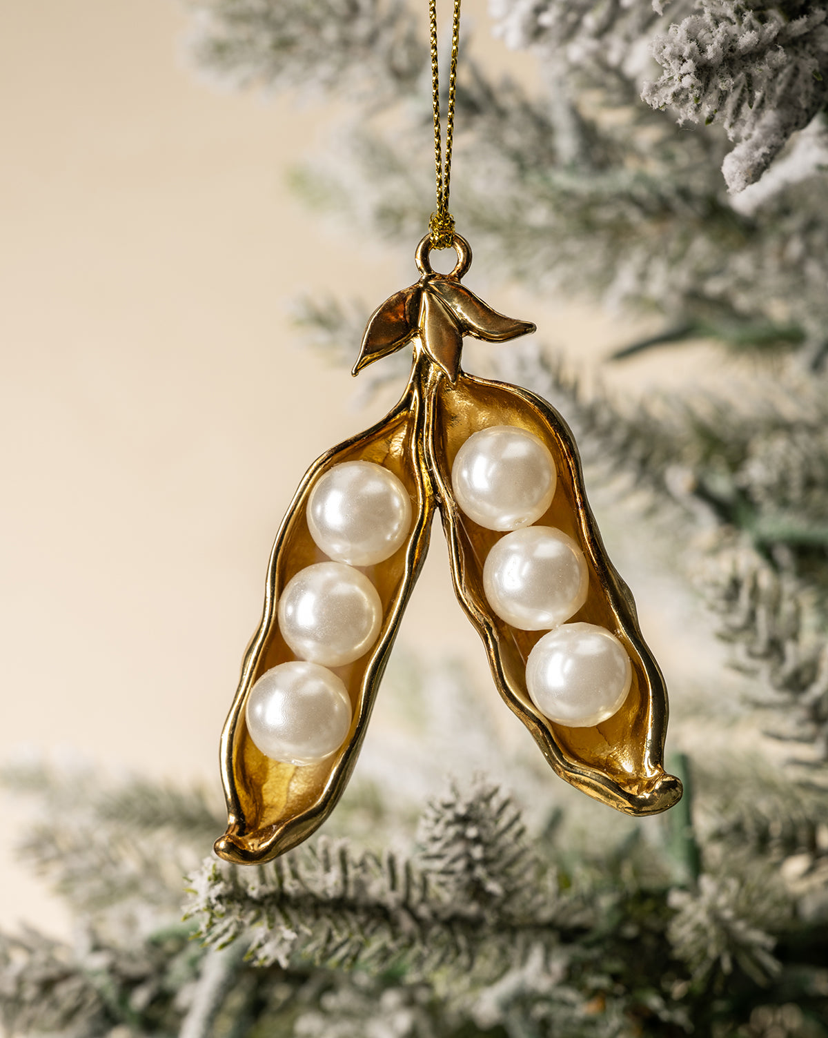The Faux Pearl Pea Pod Ornament features a gold-toned pod with six pearl-like beads inside, displayed hanging from a snow-dusted Christmas tree branch, by Vendixs