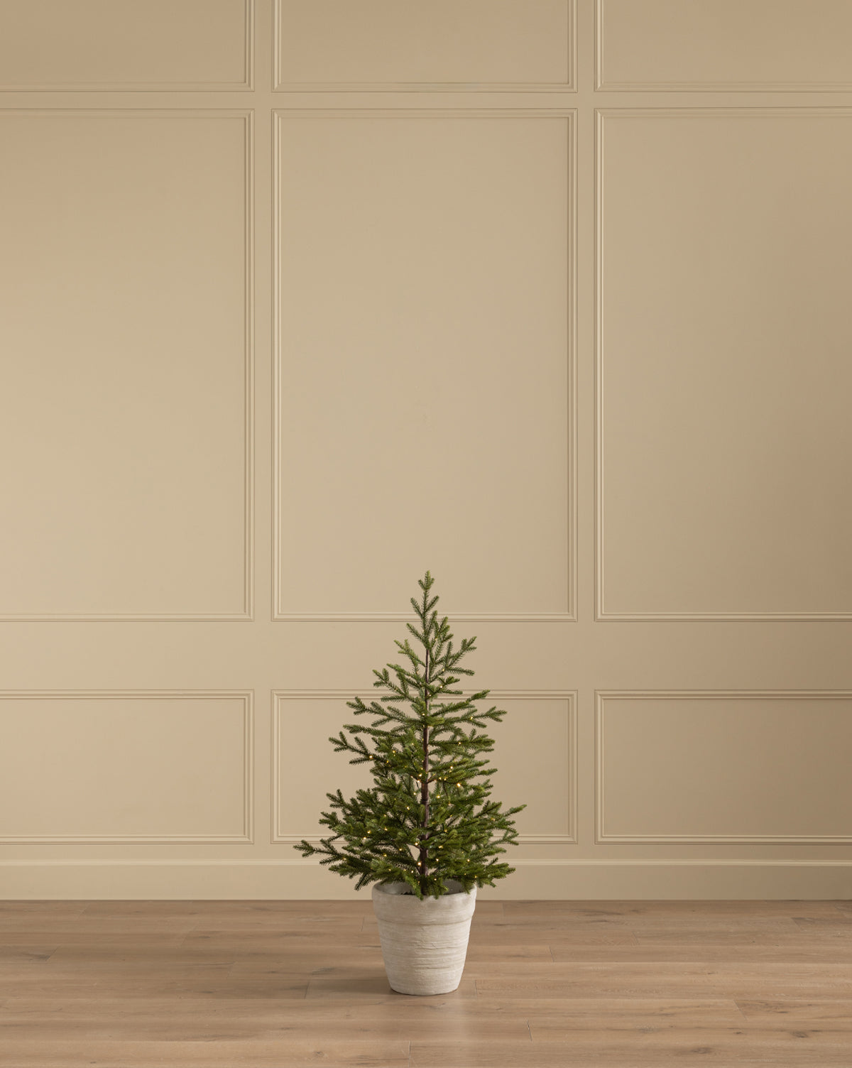 The Faux Spruce Pre-Lit Potted Tree is placed on a wooden floor before a beige paneled wall, serving as the centerpiece in a minimalist, empty room, by Vendixs
