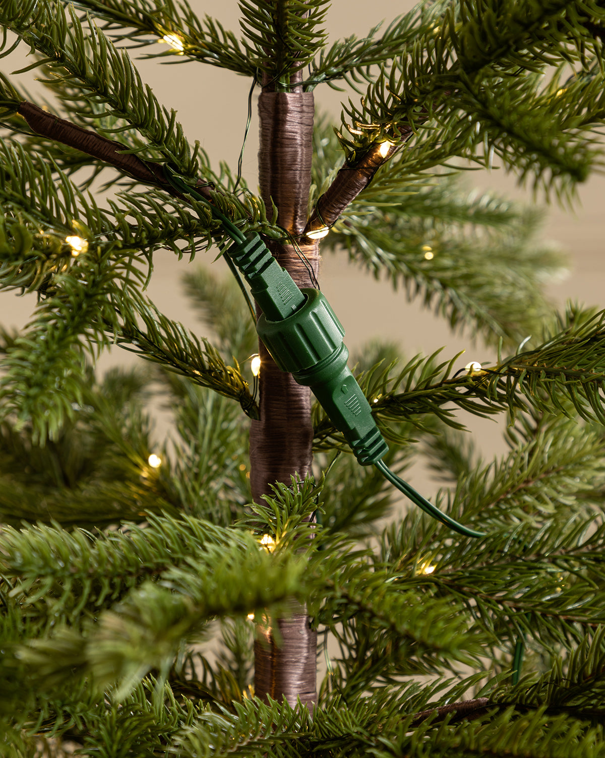 Close-up of a green electrical plug connected to a matching socket on the trunk of the Faux Spruce Pre-Lit Potted Tree, with small string lights woven among its lush branches, by Vendixs