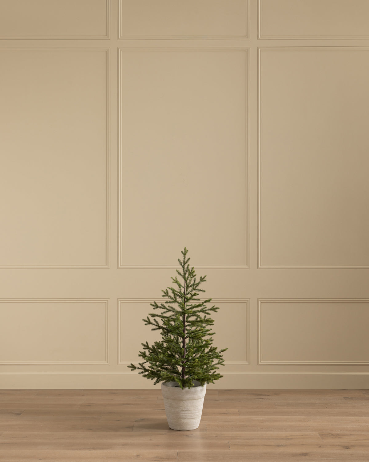 The Faux Spruce Pre-Lit Potted Tree is displayed on a wooden floor by a beige paneled wall, adding a minimalist, neutral accent to the room, by Vendixs