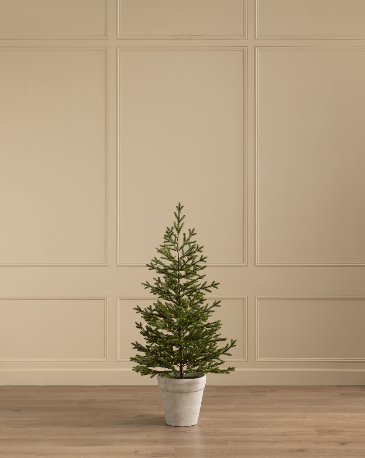 The Faux Spruce Pre-Lit Potted Tree stands on a wooden floor before a beige paneled wall, bringing greenery and soft light to the room, by Vendixs