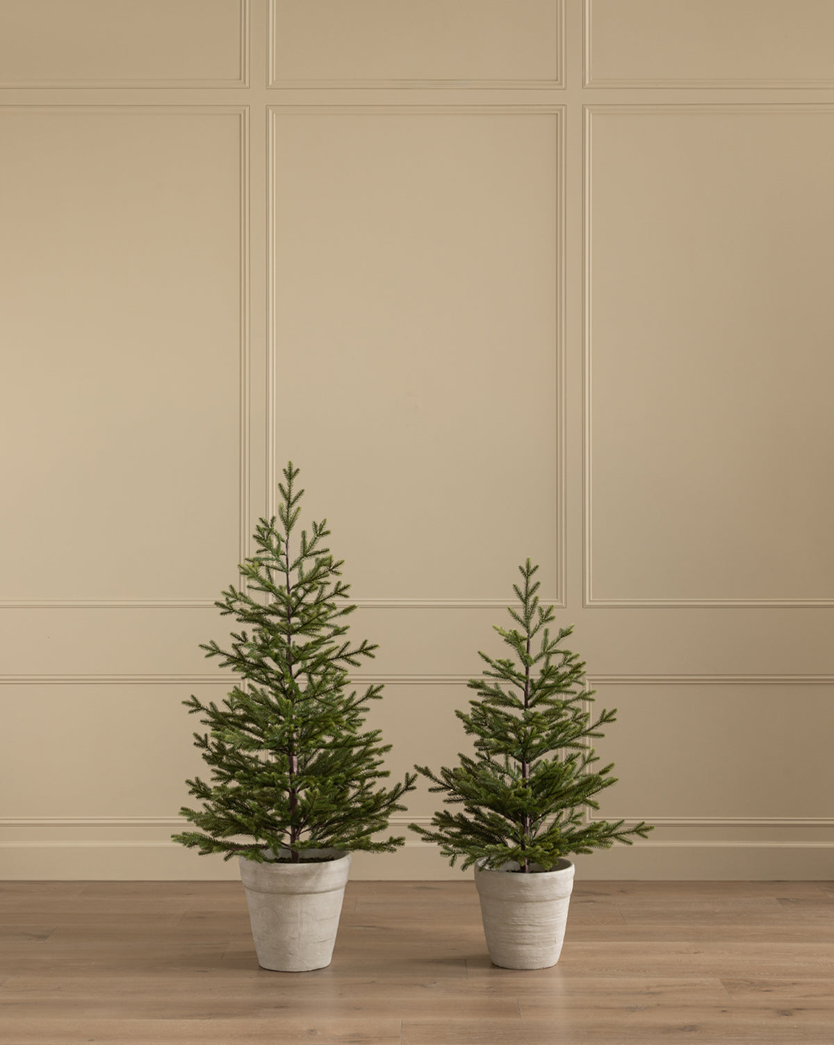 A Faux Spruce Pre-Lit Potted Tree stands on a light wooden floor beside another potted faux tree of different height, set against a beige paneled wall, by Vendixs