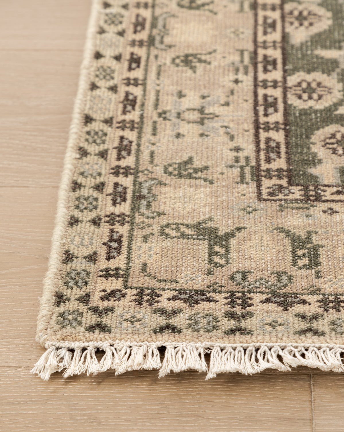 A close-up of the Fernwood Hand-Knotted Wool Rug shows its beige and green wool, fringed edges, and geometric floral motifs, elegantly placed on a light wooden floor, by Vendixs