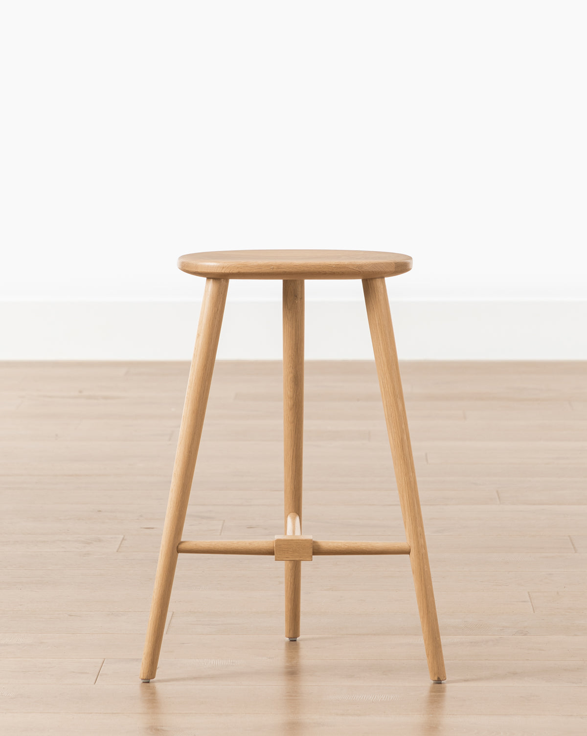 The Fielder Counter Stool features a round seat and three legs in a light oak finish, shown on a light wood floor beside a white wall, by Vendixs