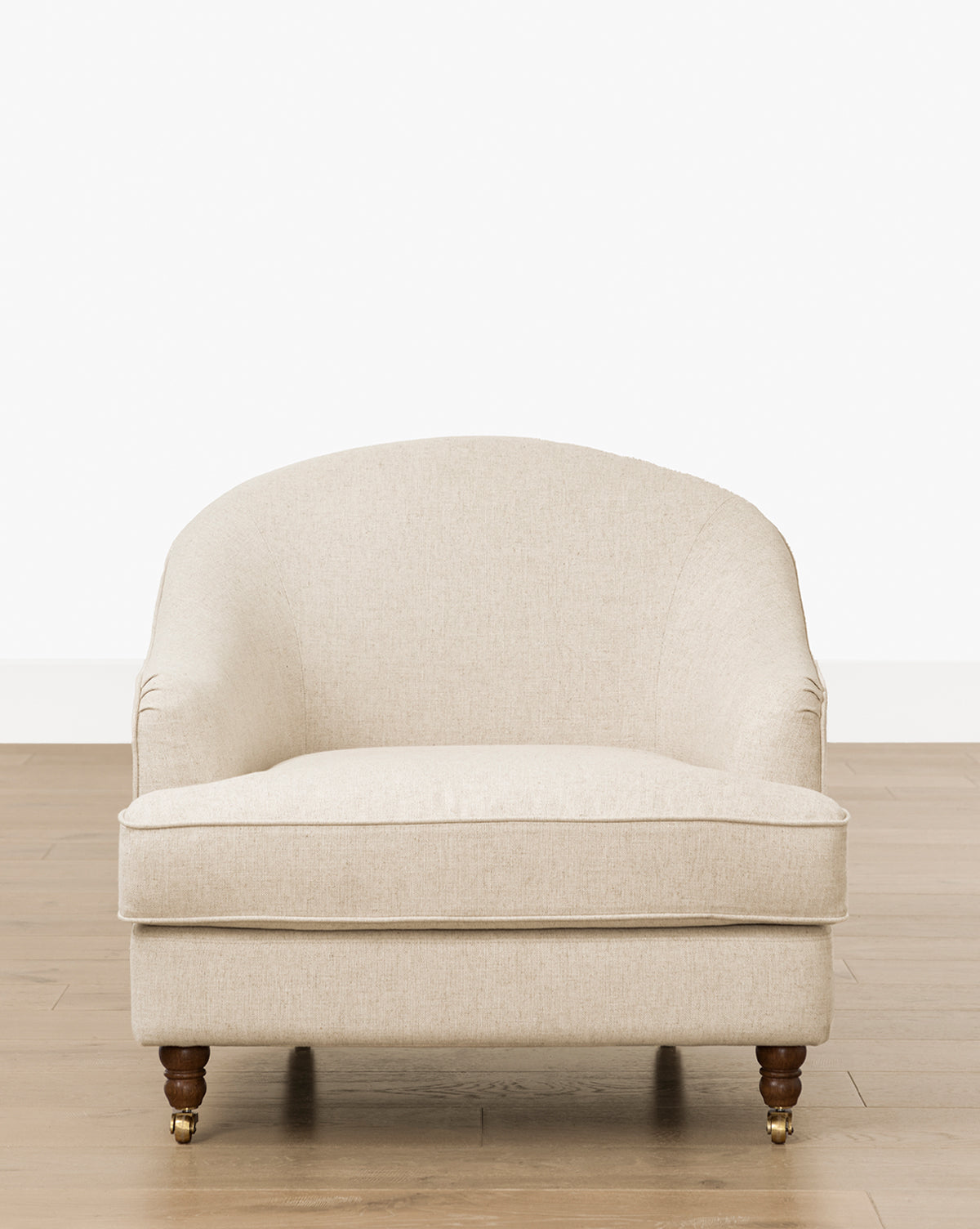 The Makerspalm Fletcher Lounge Chair (Ready to Ship) is a beige upholstered armchair with a rounded back, cushioned seat, and wooden legs with brass casters, shown on a light wood floor against a plain white background by Vendixs