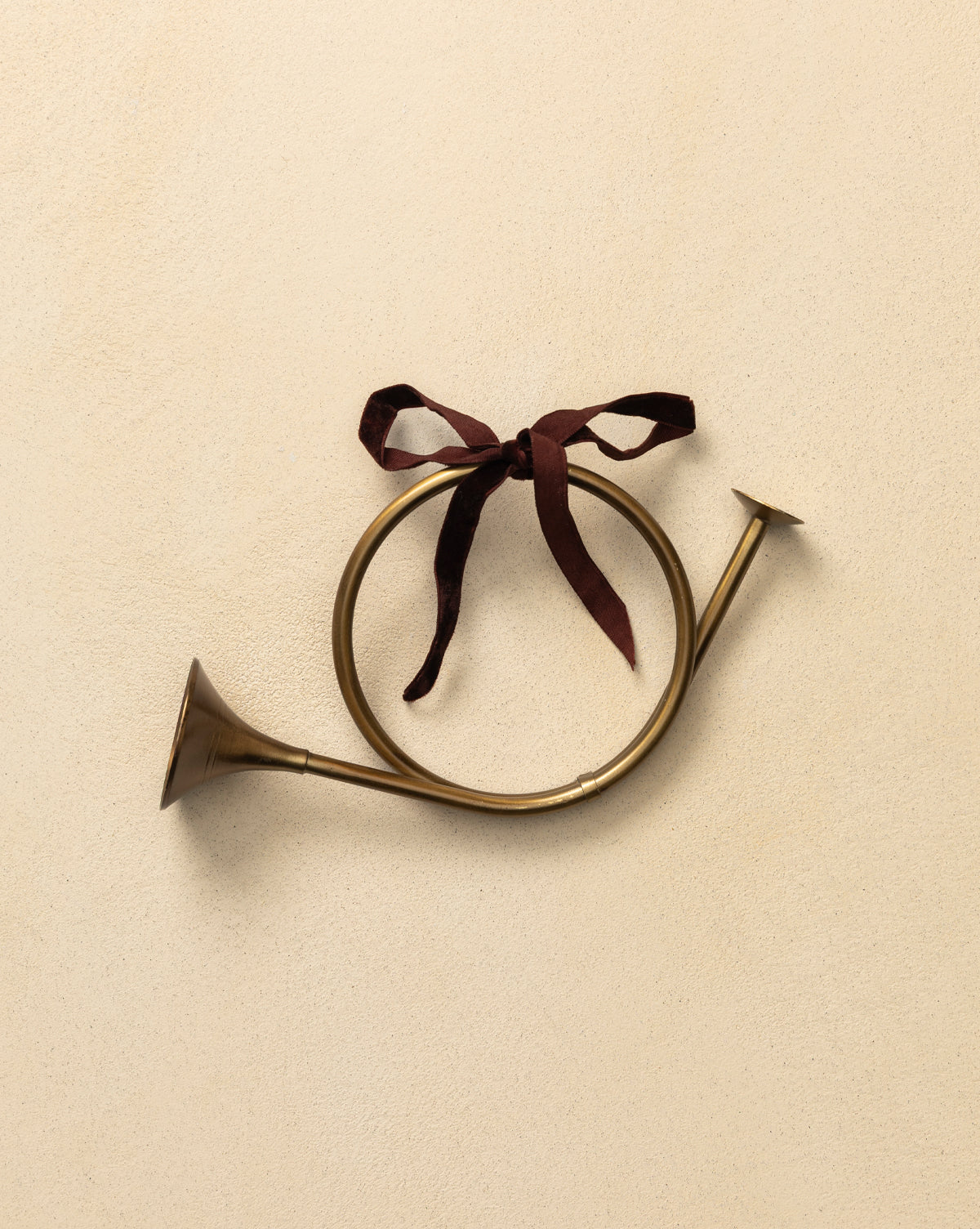 The French Horn Ornament features a brass finish and a maroon ribbon bow, hanging on a light beige textured wall—an ideal touch to enhance your holiday tree décor, by Vendixs